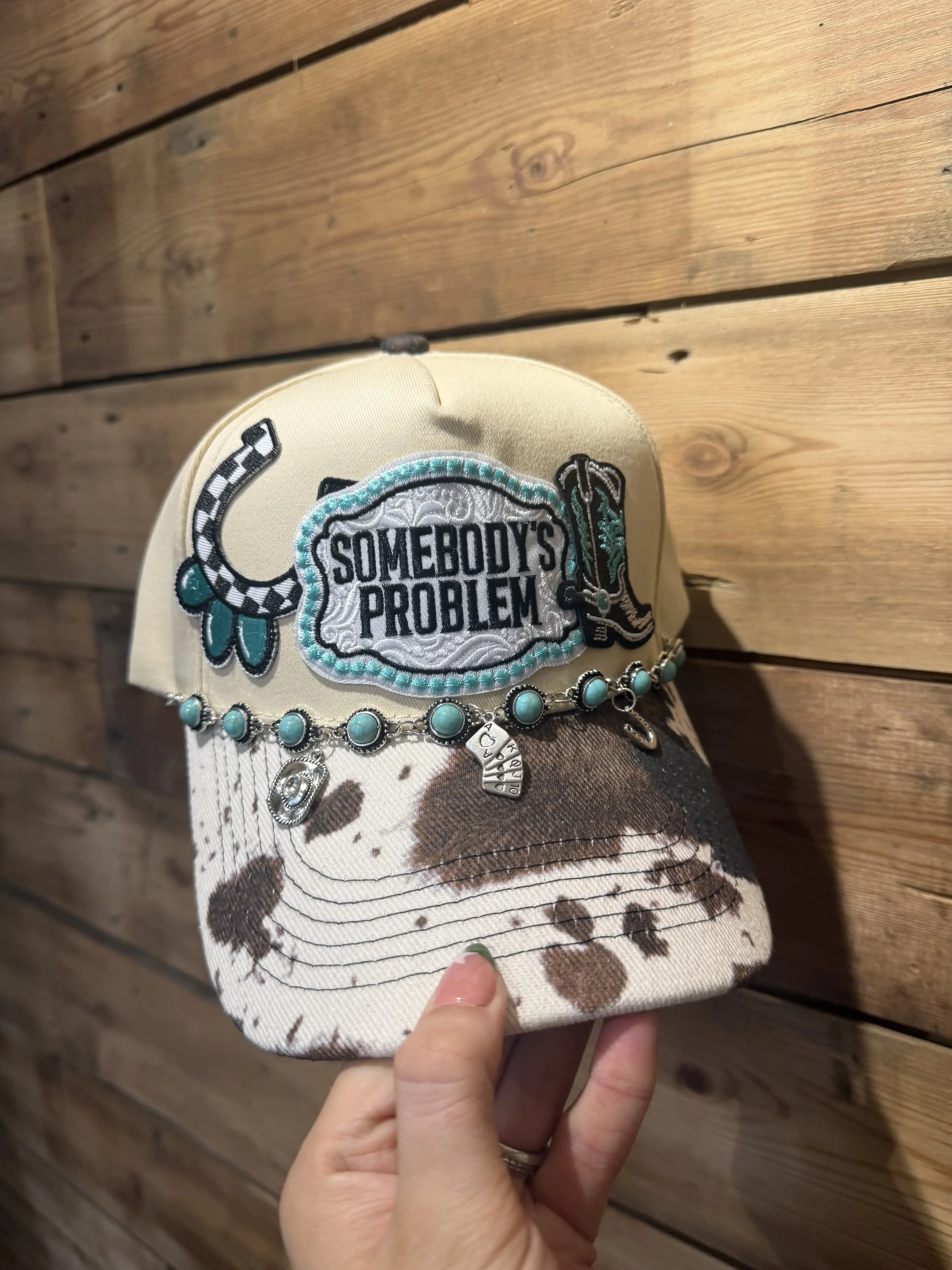 A hand holding a baseball cap with embroidered patches that say 'Somebody's Problem', a horseshoe, and a cowboy boot. The cap is resting on a wood paneled wall.