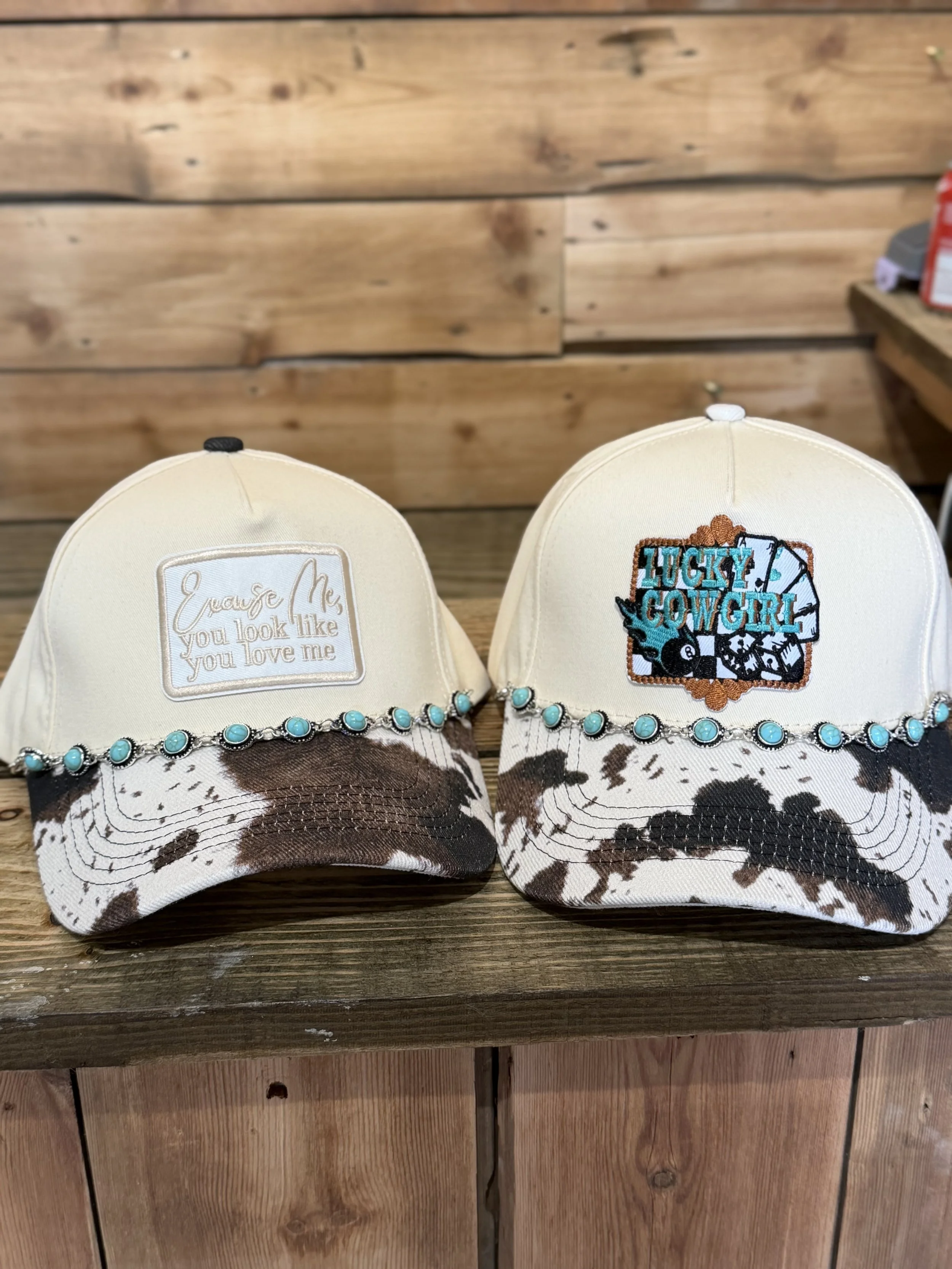 Two baseball caps with cowhide pattern brims and decorative turquoise beads, placed on a wooden surface. The cap on the left has a patch with the text 'Then you look like you love me,' and the cap on the right has an embroidered patch reading 'Lucky 