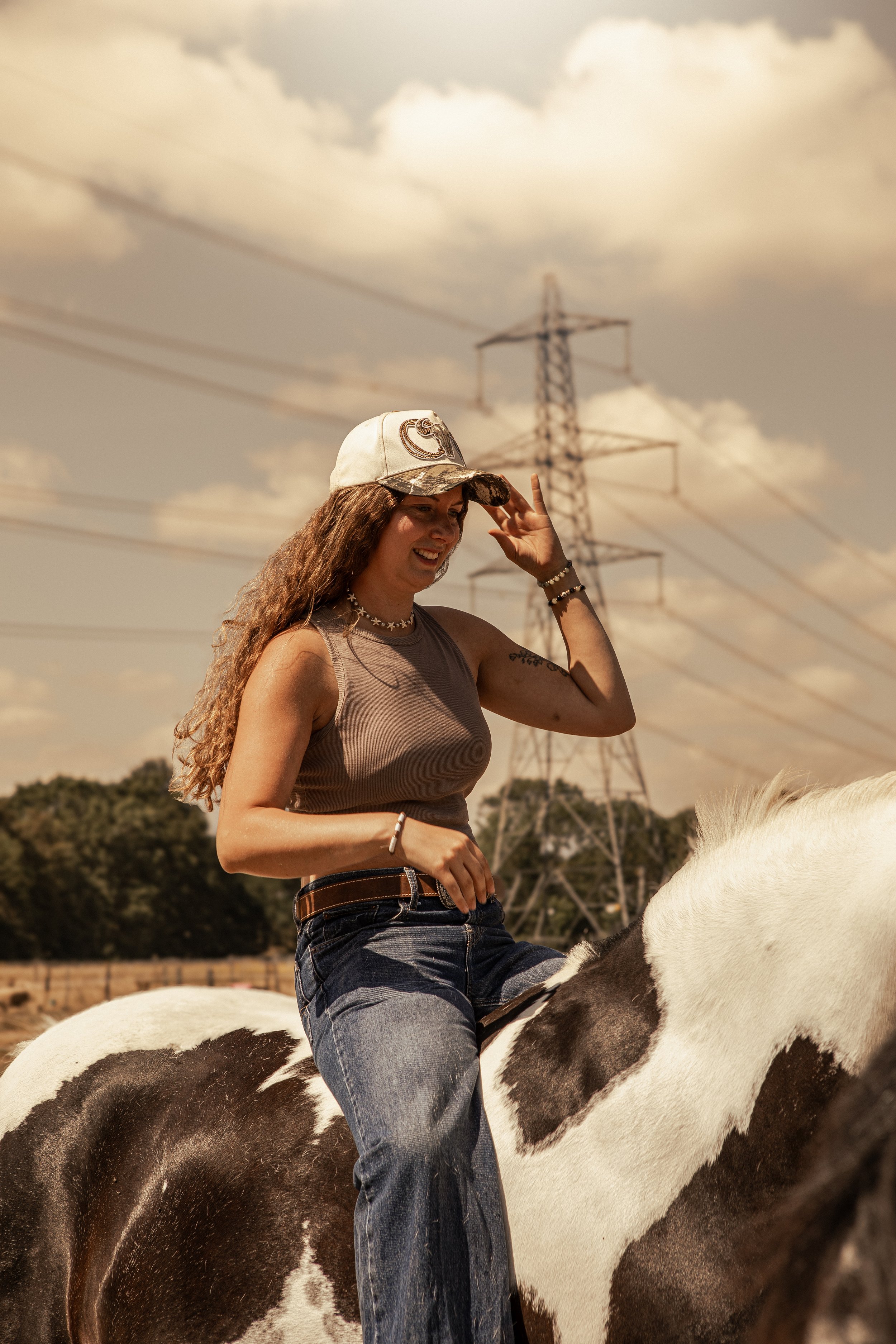 Woman with long curly hair wearing a white cap, gray tank top, and dark jeans, sitting on a black and white horse with a saddle, smiling during daytime with a cloudy sky and power lines in the background.