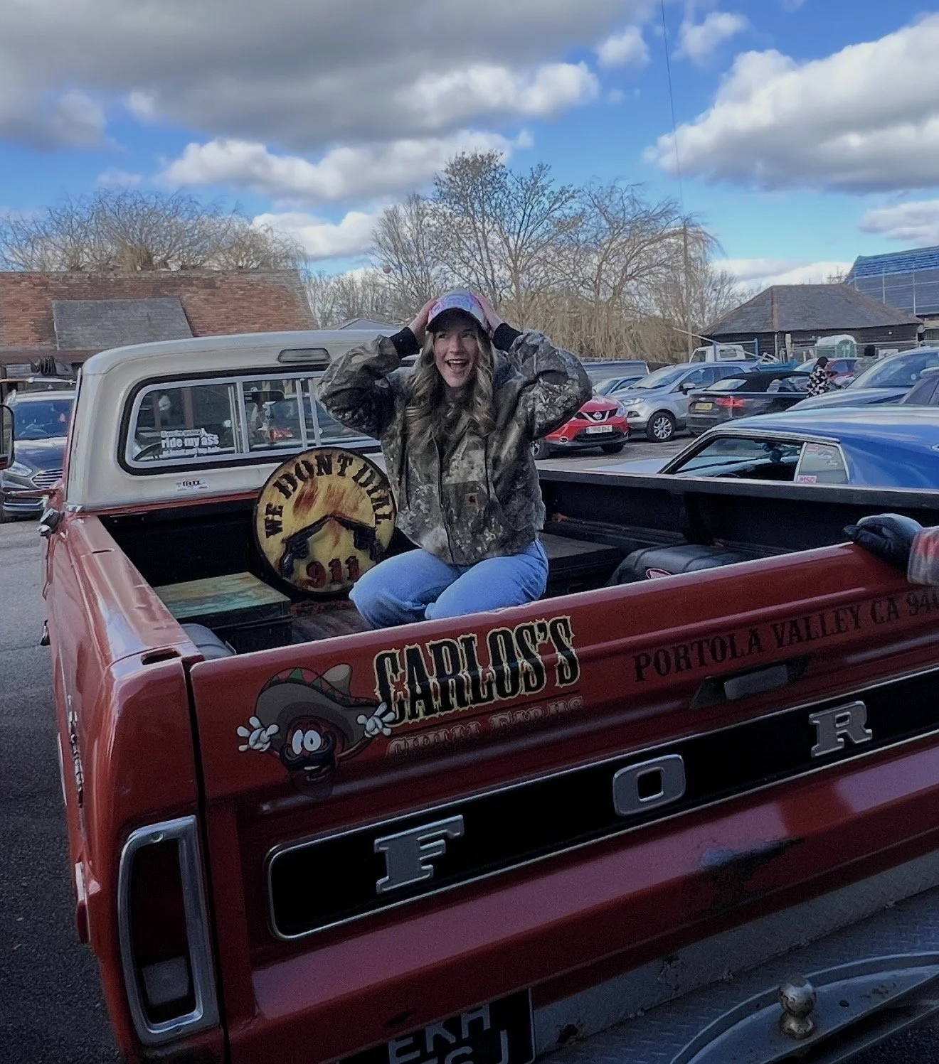 A woman sitting in the back of a red Ford pickup truck, smiling and holding her hands on her head, wearing a camo jacket, blue jeans, and a cap. The truck has a sticker that says 'We Hunt Wildlife 911' and other decals.