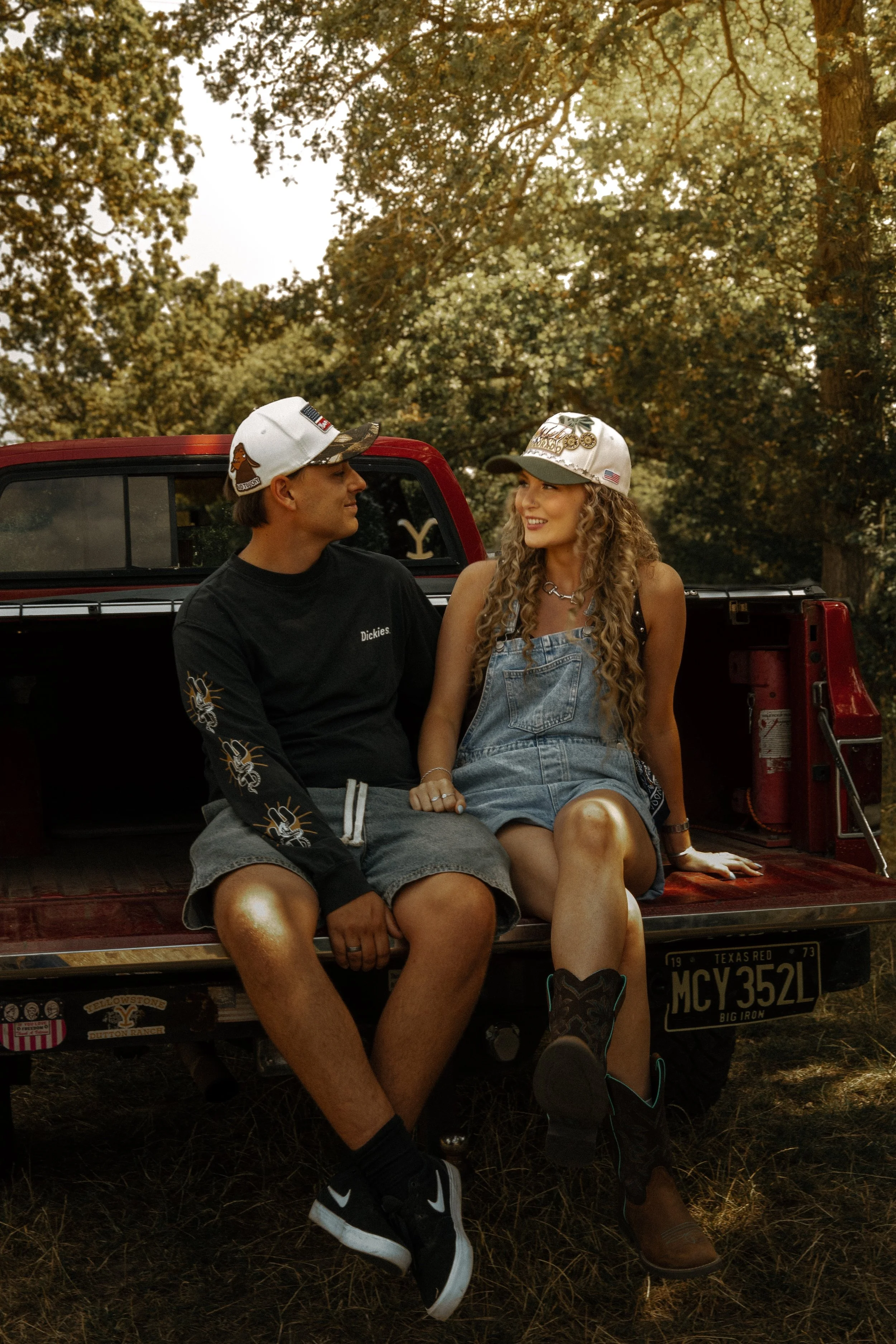 A young man and woman sitting on the tailgate of a red pickup truck in a wooded outdoor setting, wearing casual clothing and hats, with the woman smiling at the man. The truck has various stickers and a Texas license plate.