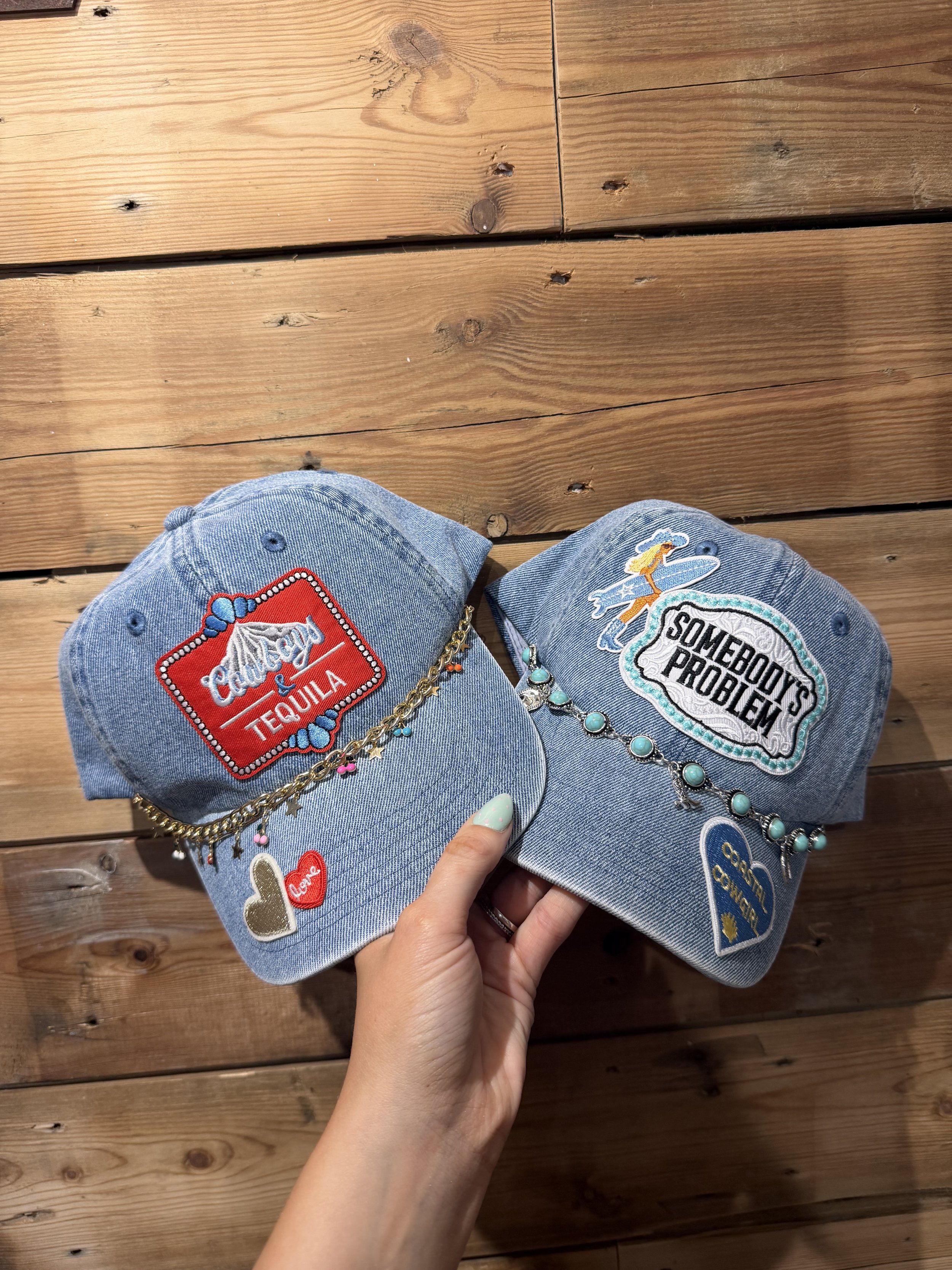 Two denim baseball caps decorated with patches and jewelry, held in a person's hand over a wooden surface.