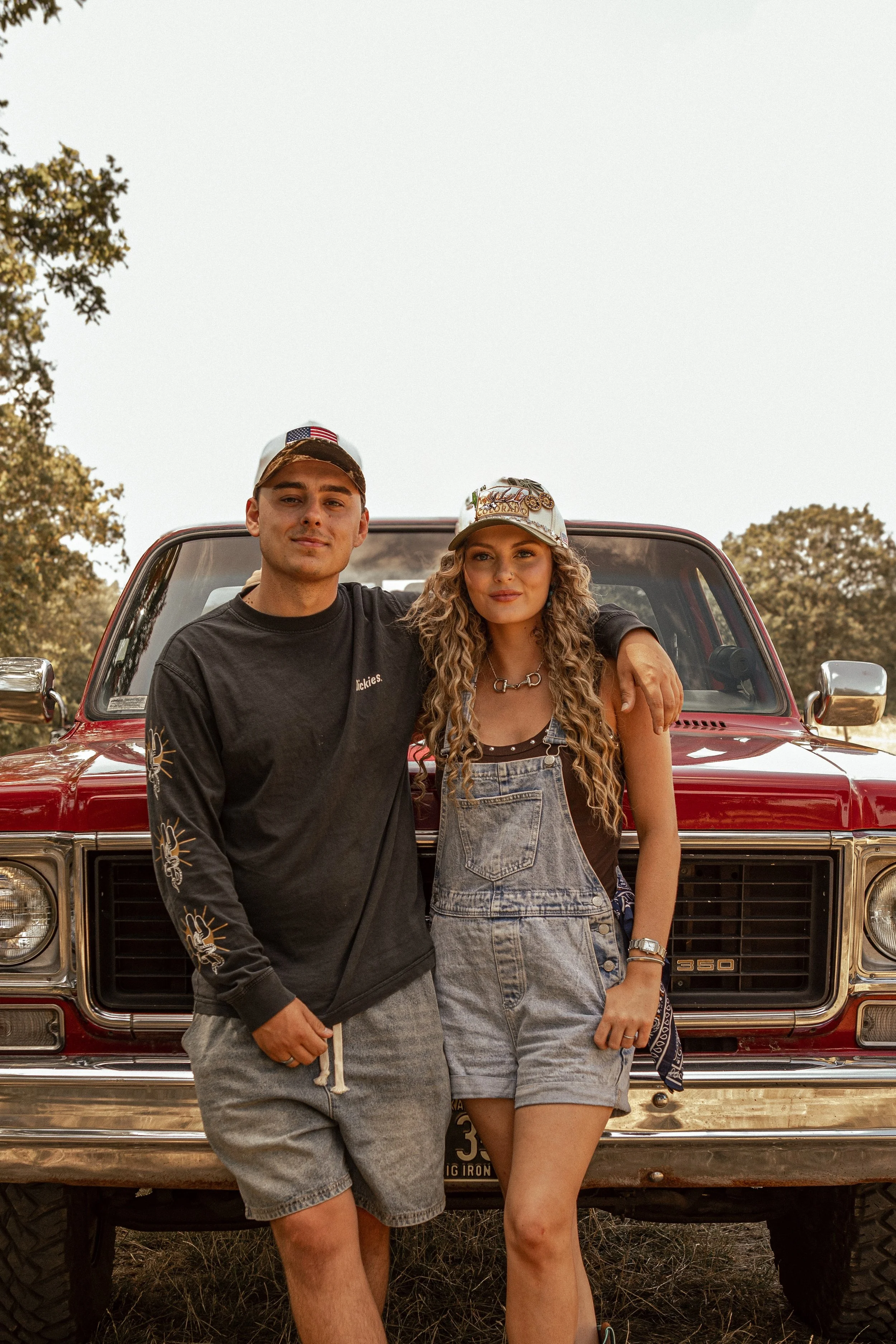 A young man and woman stand in front of a red pickup truck, with trees and a clear sky in the background. The man wears a camo hat with an American flag patch and a black long-sleeve shirt. The woman has curly hair, a colorful hat with patches, and denim overalls. They are outdoors during sunset or late afternoon.