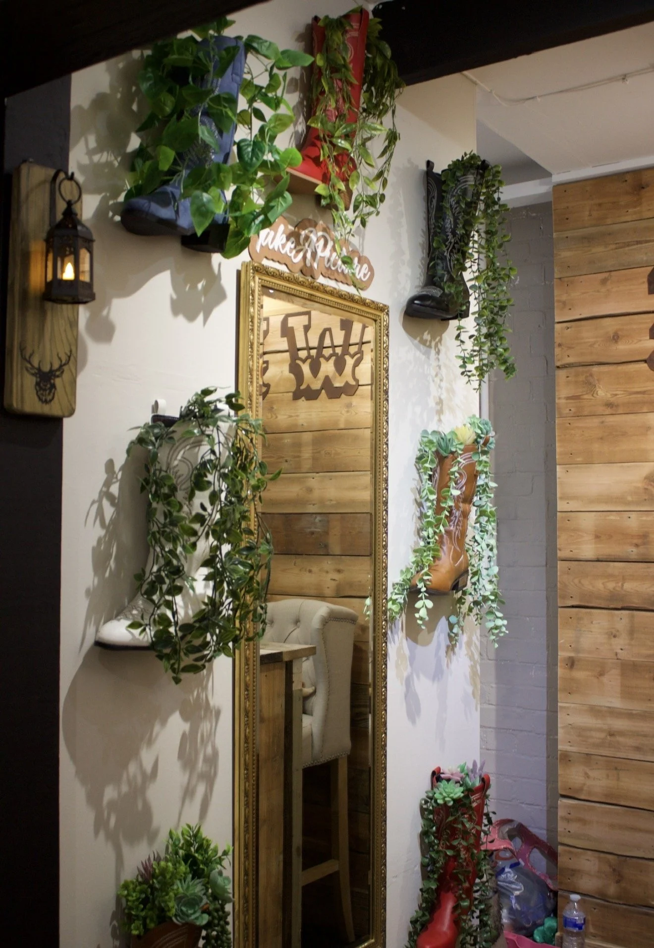 Decorated wall with hanging plants, a mirror, a small shelf, a wooden cowboy boot with plants inside, and a sign that says 'Take a Minute.'