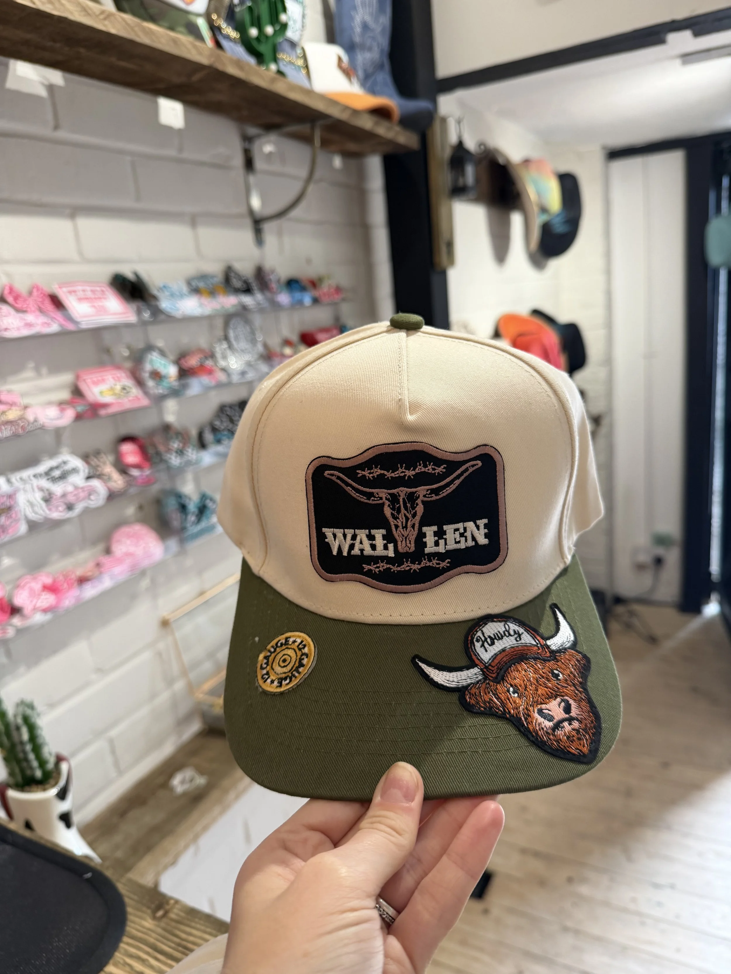 Hand holding a cap with a 'Wild One' patch and a tiger embroidery, in a retail store with other hats and accessories on display.