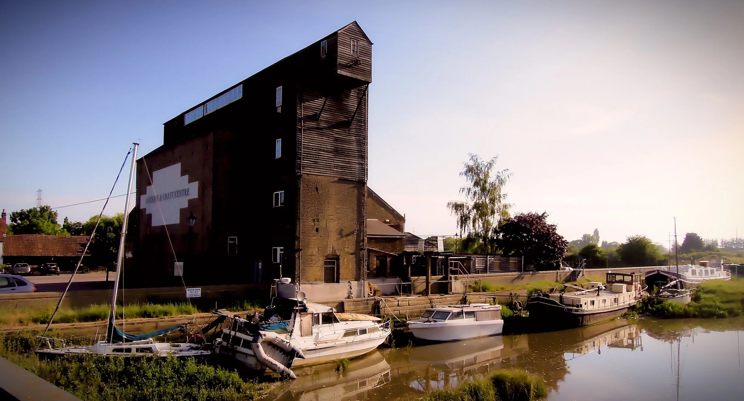 A large, old brick building next to a small harbor with several boats docked. The building has a weathered wooden extension at the top and a sign reading 'MOBILE CRAFT CENTRE'. There are trees and a partly cloudy sky in the background.