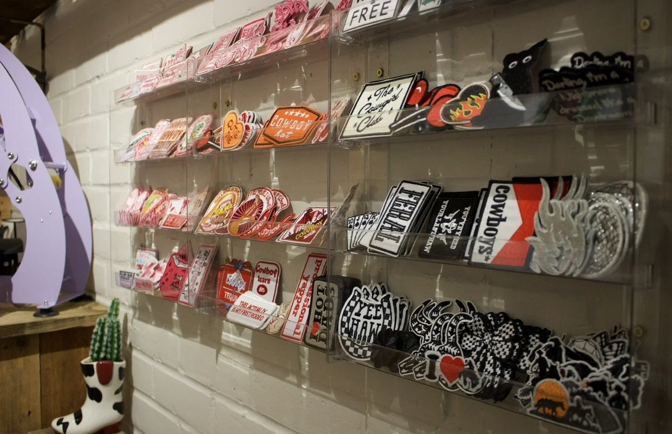 Clear acrylic wall-mounted display case with various patches and embroidered pins, featuring themes like cowboys, flames, and black and white patterns, against a white brick wall in a boutique or store setting.