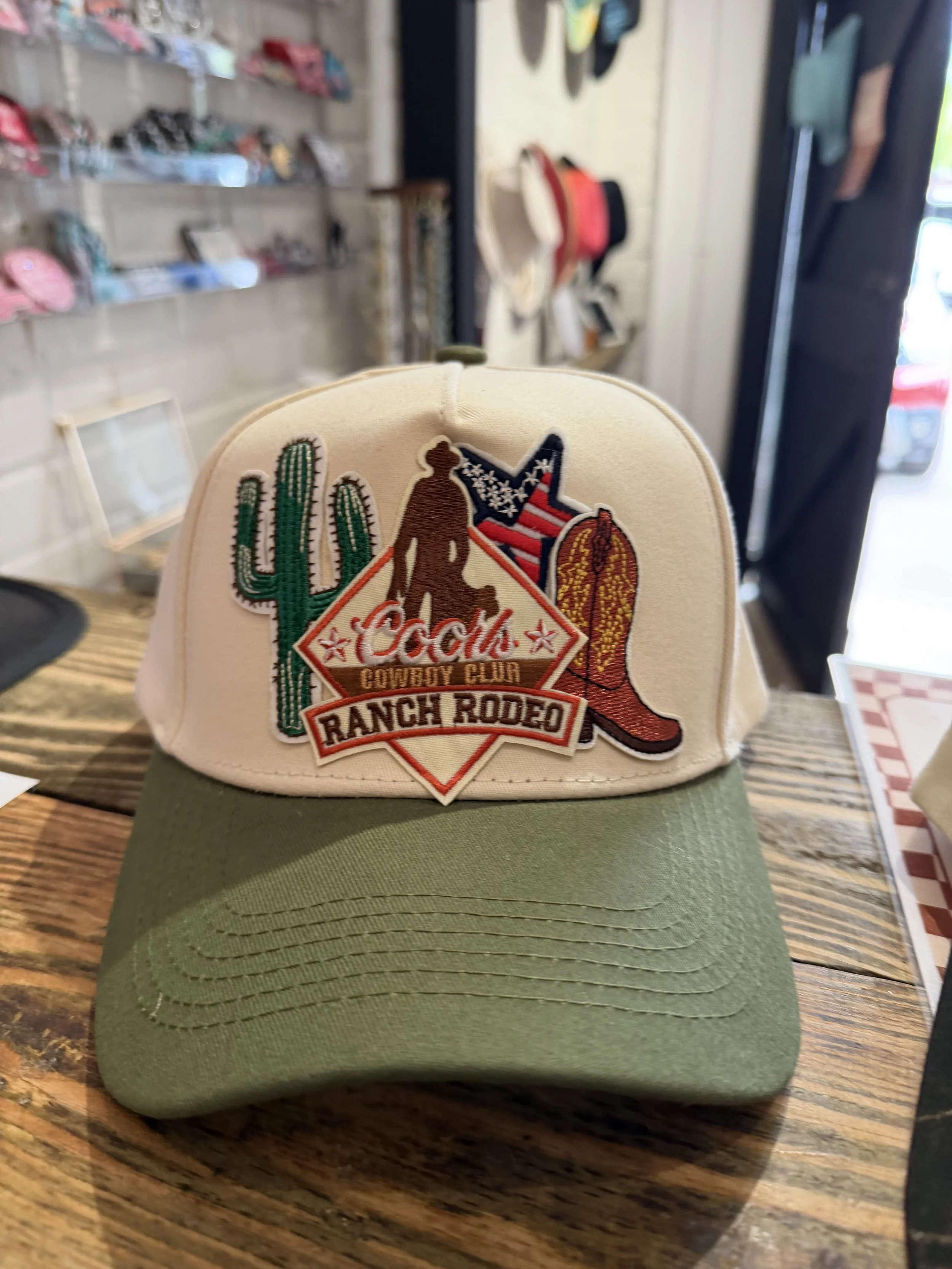 A beige cap with a patch reading 'Coca-Cola Cowboy Club Ranch Rodeo' featuring a cowboy hat, cactus, and an American flag design, placed on a green surface in a store with hats and accessories in the background.