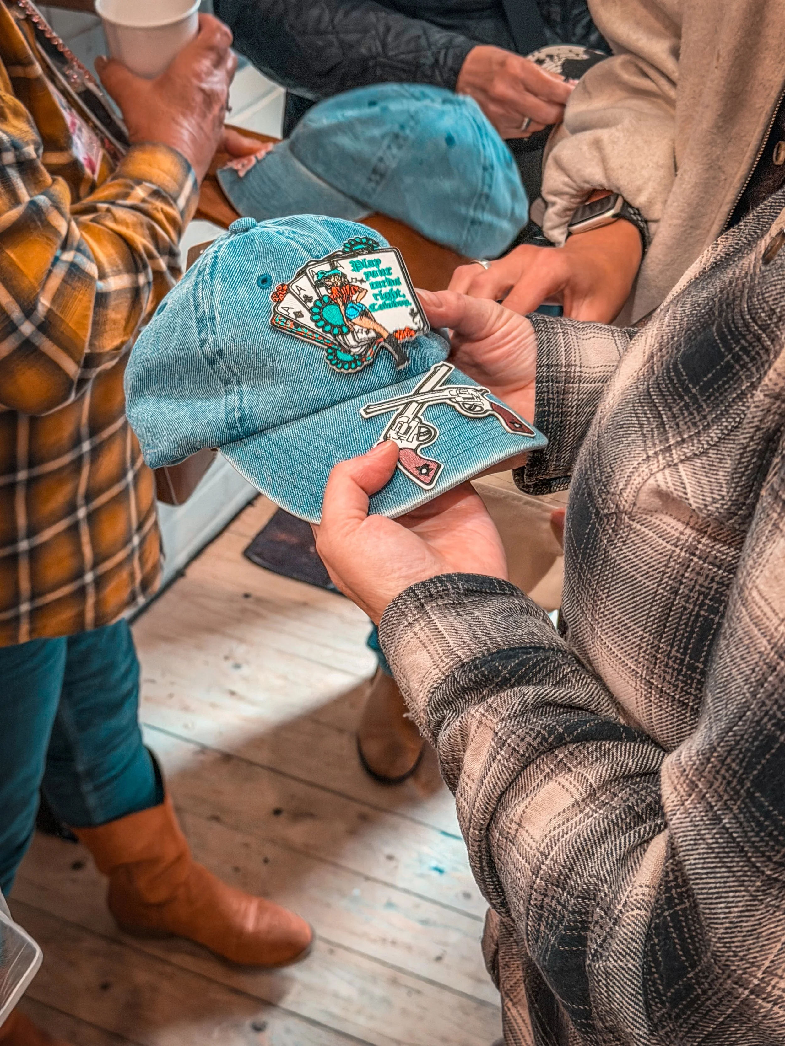 Group of people gathered around a table, holding and displaying various patches and stickers, with some holding cups. Focus on garments like a denim cap with patches, plaid shirt, and cozy jackets. Wooden floor is visible.