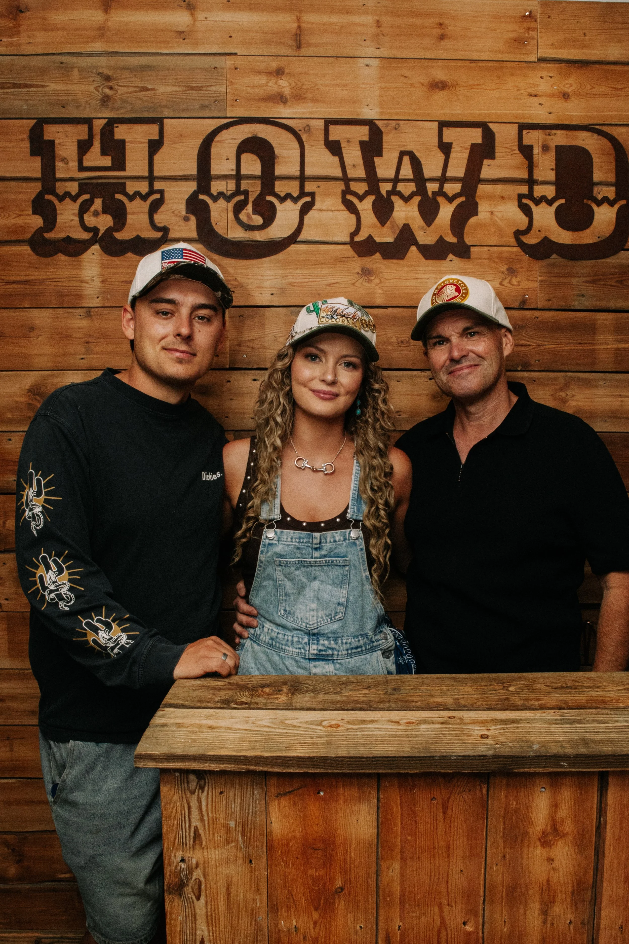 Three people standing in front of a wooden wall with a large sign that says 'COWBOY'. They are wearing casual clothing and hats, and are posing for the photo.
