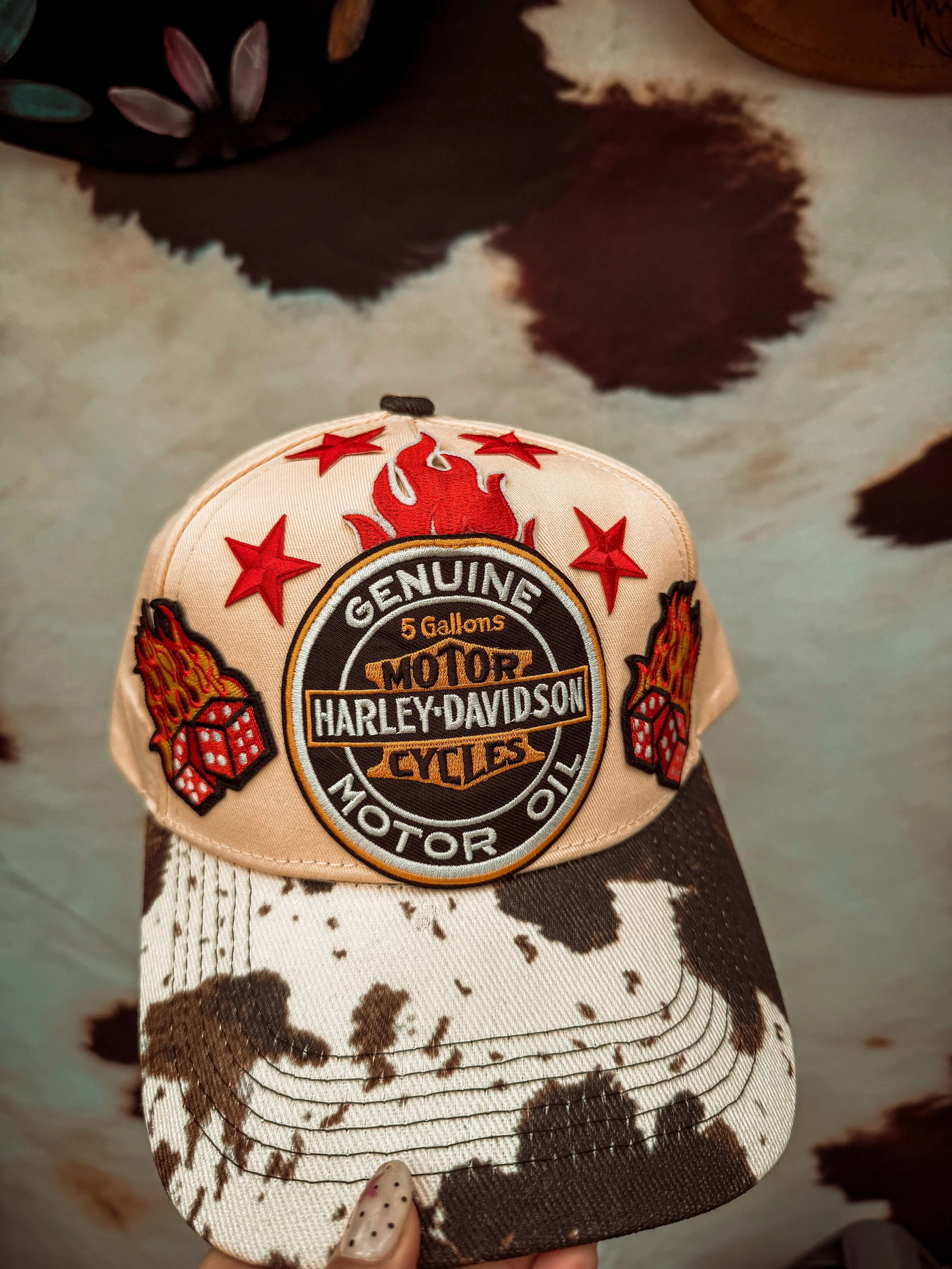 A beige Harley-Davidson baseball cap with red stars and flames embroidery on the front, resting on a cowprint fabric background with black, white, and brown spots.