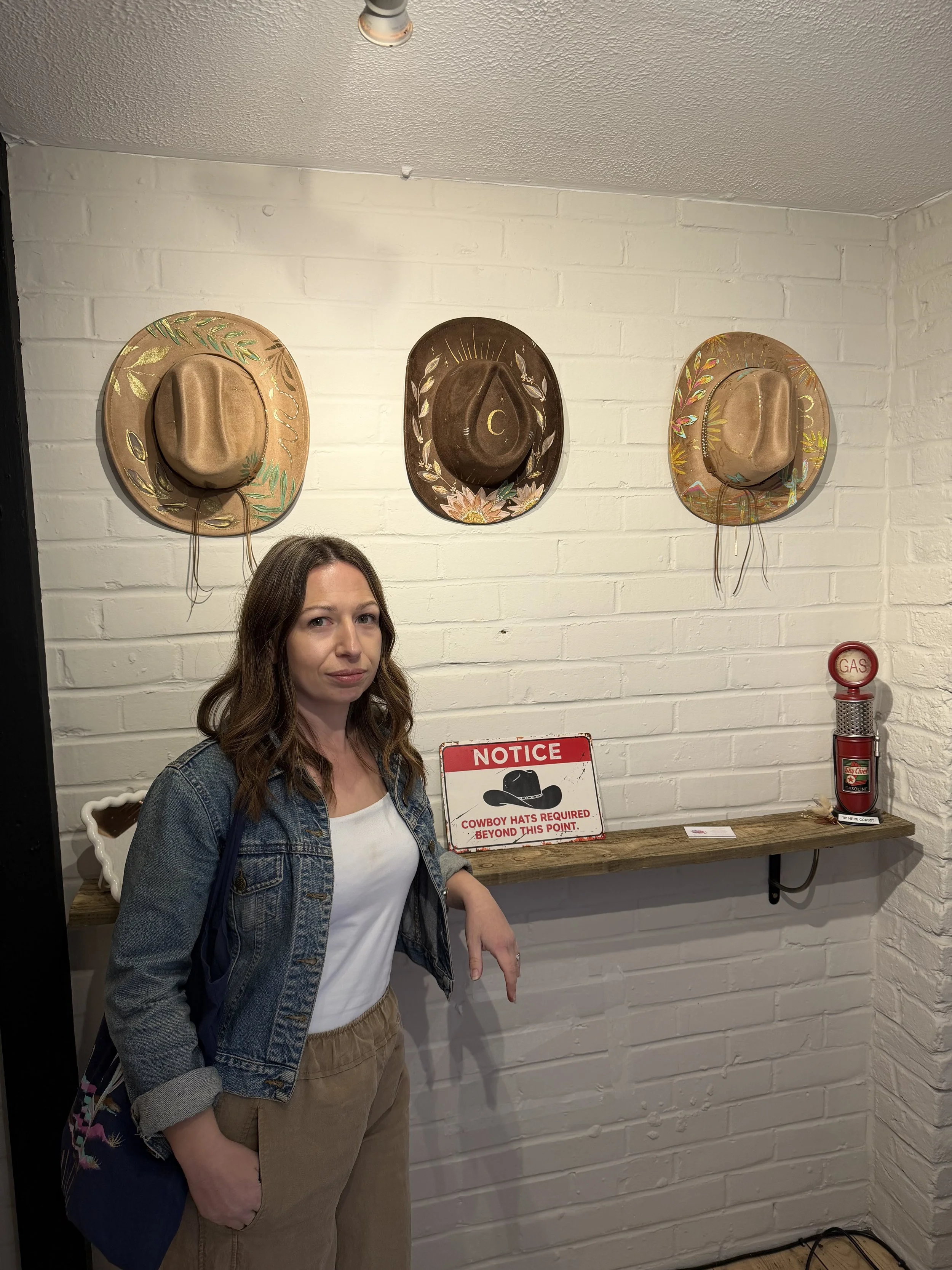 A woman with long brown hair, wearing a denim jacket and khaki pants, stands in front of a white brick wall with three decorative cowboy hats hanging above her. There is a red and white sign that says 'NOTICE COWBOY HATS REQUIRED BEYOND THIS POINT' and a vintage-style fire extinguisher on a wooden shelf to her right.