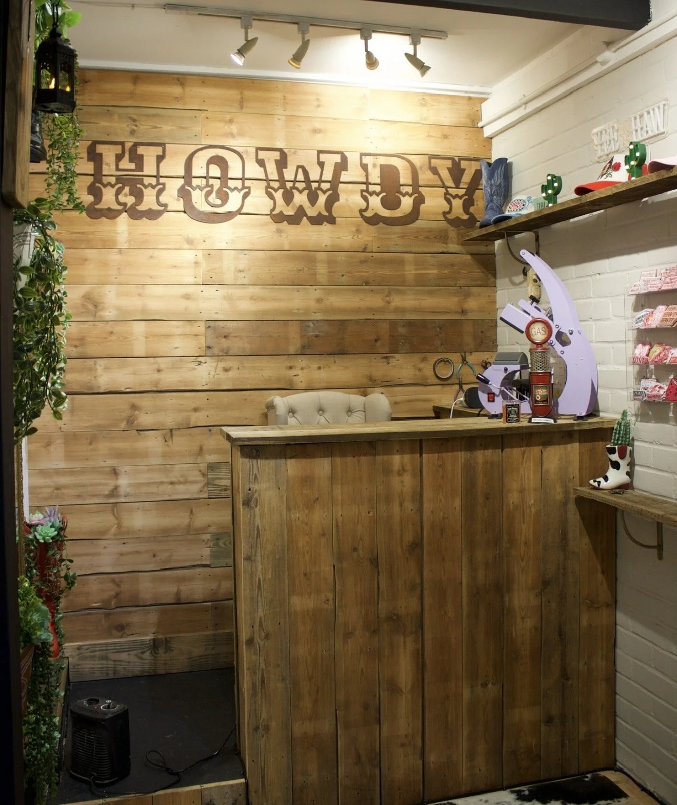 Indoor scene with a wooden counter, a vintage cash register, and decorative cowboy boot with cactus design. Wooden wall with a sign that says 'Howdy' in metal letters. Small shelf with cowboy hat, cactus decor, and other items. White brick wall with shelving and plants to the left. Black heater at the bottom.