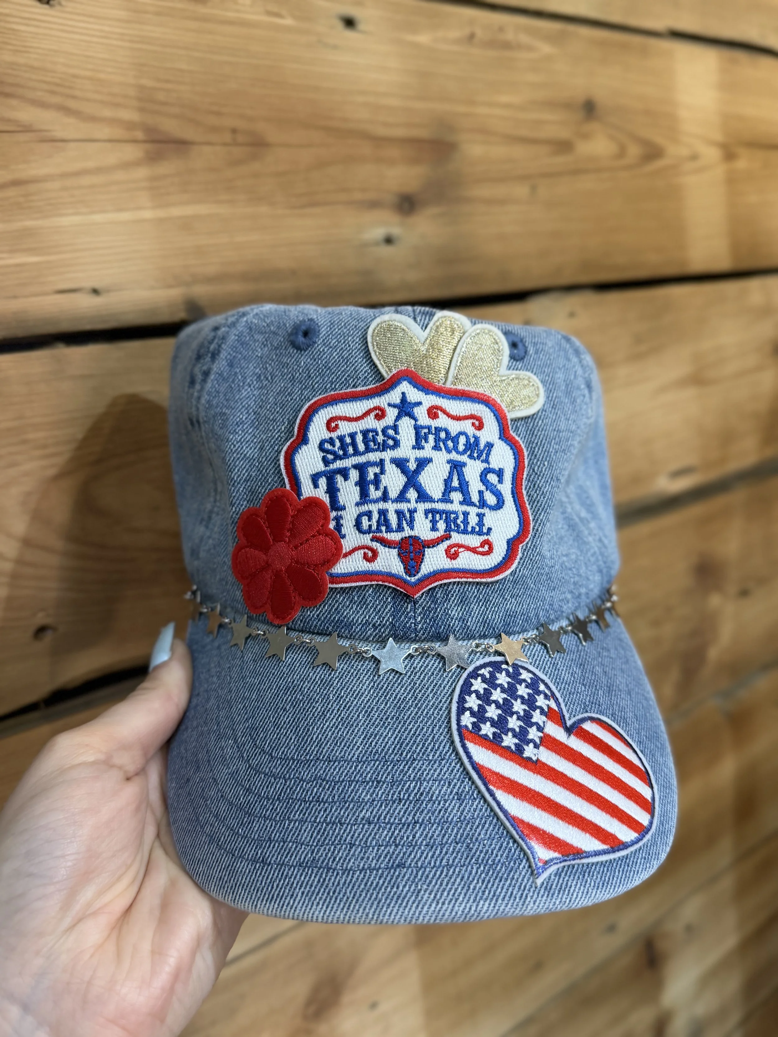 A denim cap with patches and embroidered text reading 'She’s from Texas she can’t feel,' with star and heart decorations, held by a person's hand against a wooden background.