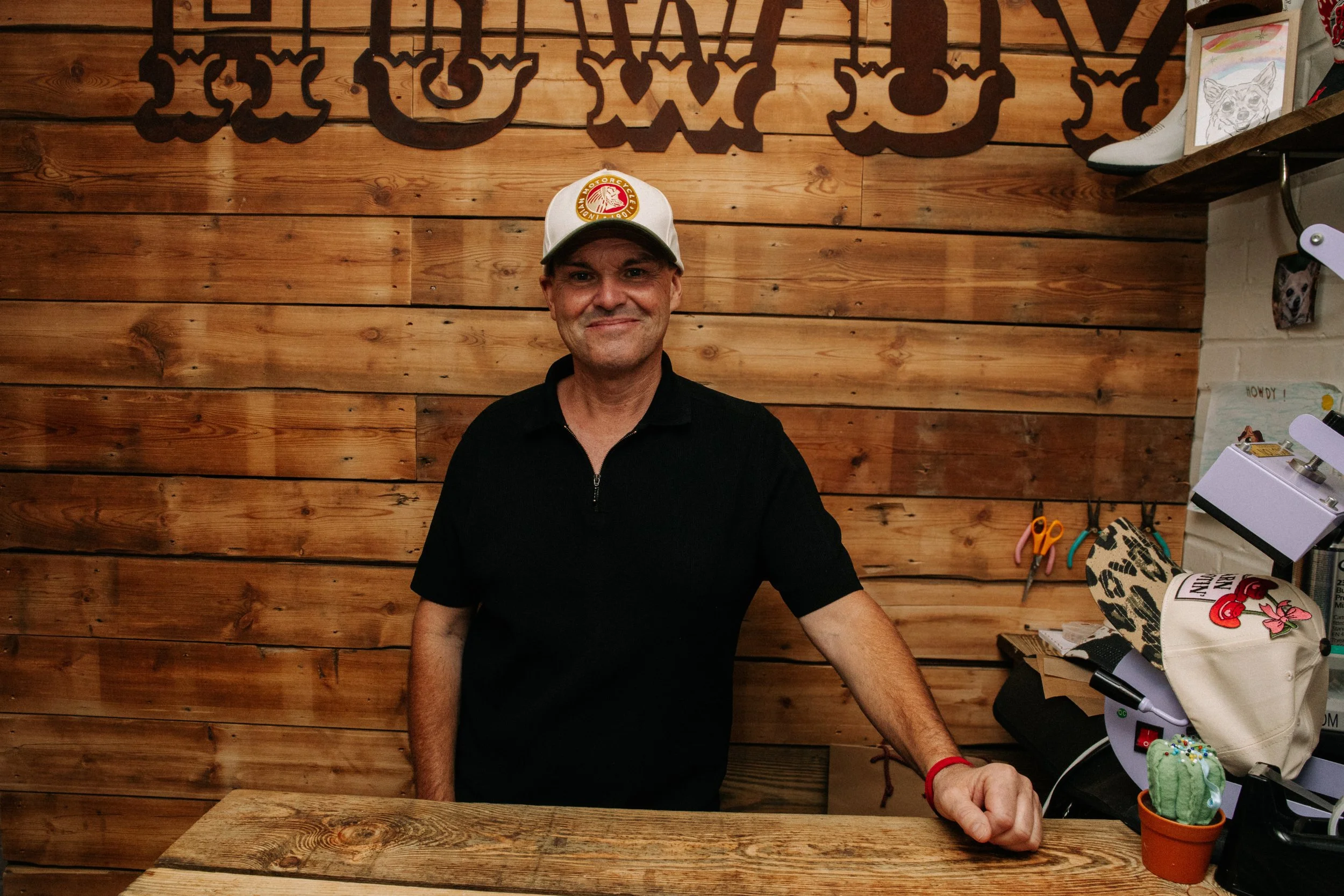 A man wearing a white cap with a red and yellow logo stands in front of a wooden wall with large decorative lettering. He is smiling and leaning on a wooden table with crafting supplies, including a potted cactus, scissors, a heat press, and fabric items.