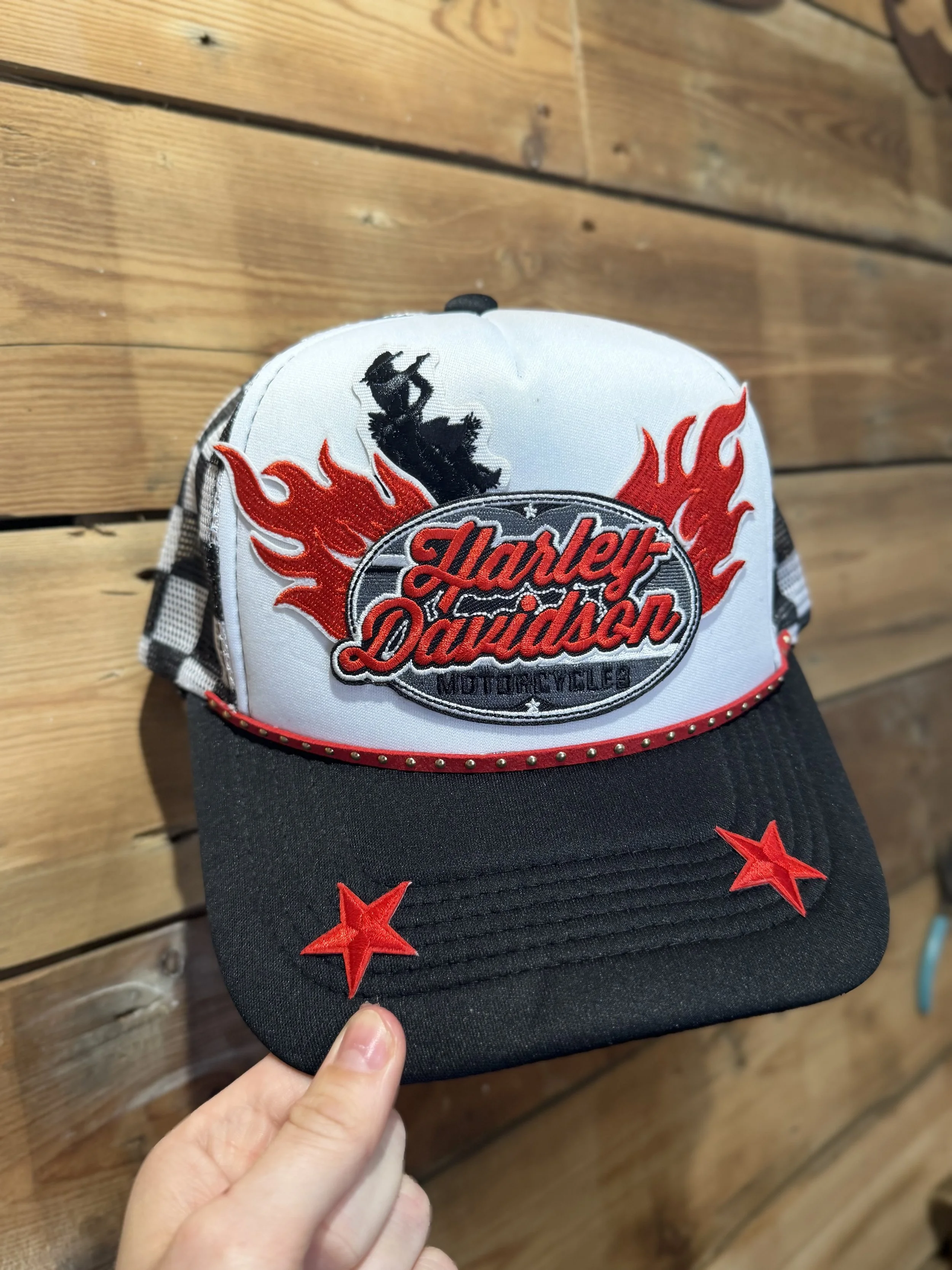 A person's hand holding a Harley Davidson cap with black, red, and gray decorations, featuring a black motorcycle silhouette and red flames on the front, and red stars on the black brim, against a wooden background.