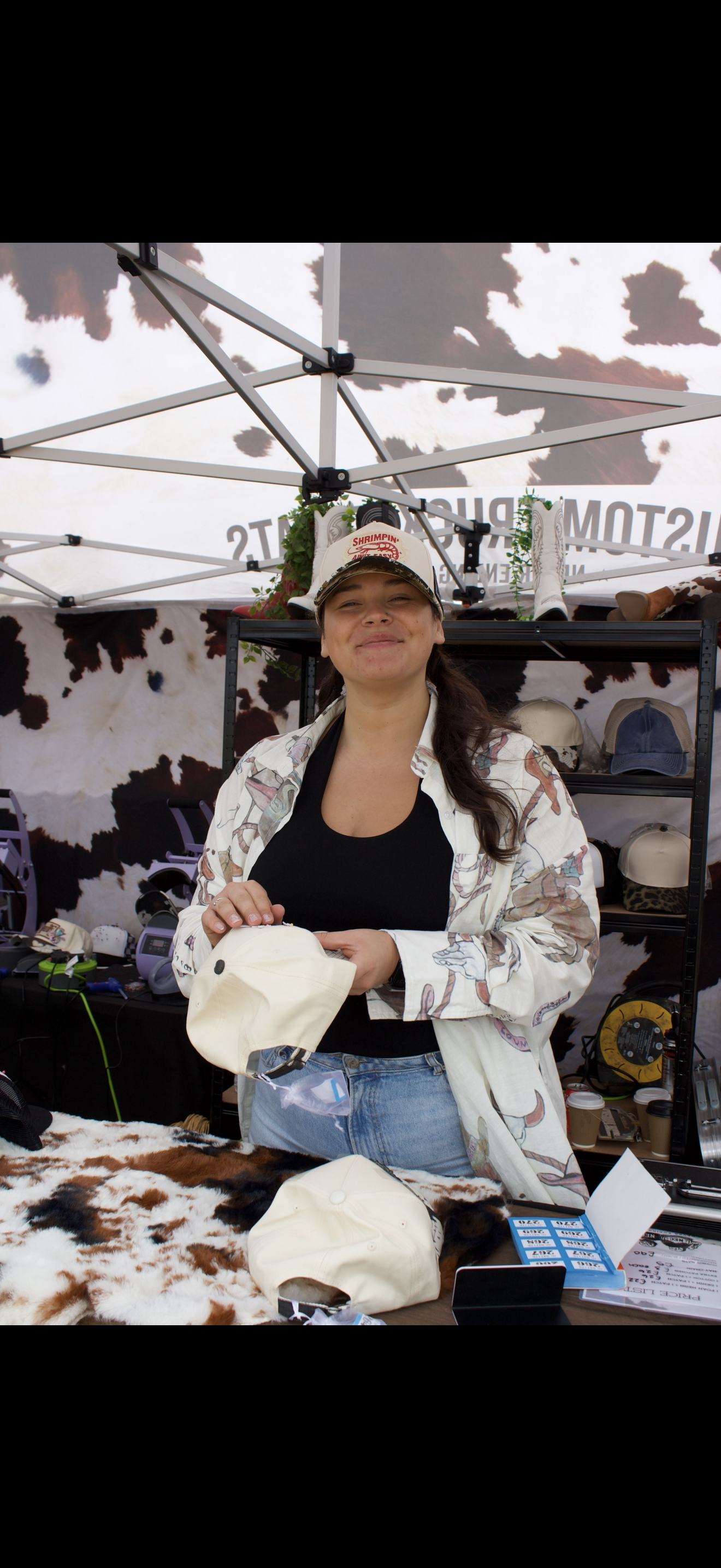 A woman standing at a booth selling hats, with shelves of hats and equipment behind her, under a cow-print canopy.