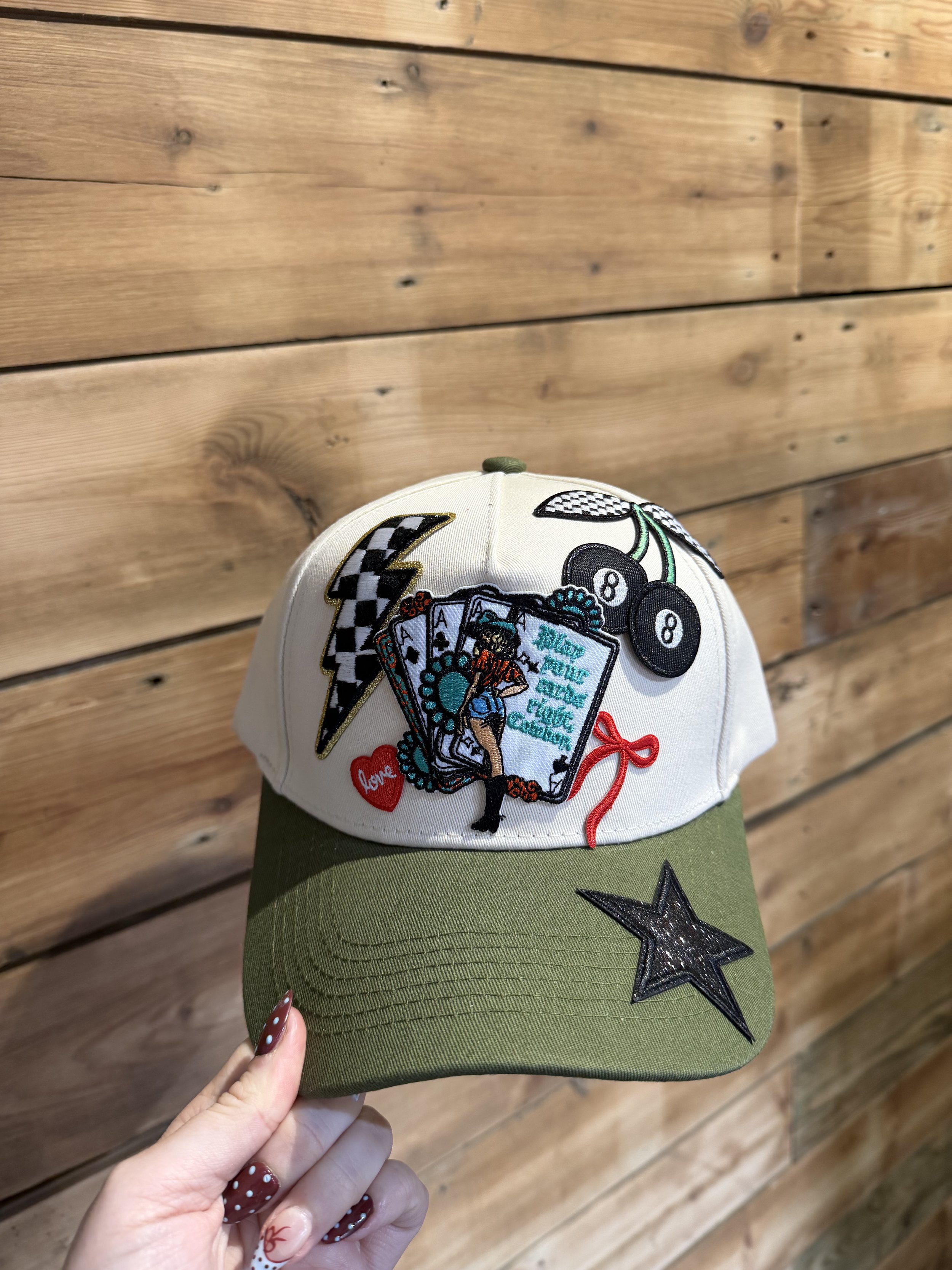 A hand holding a baseball cap with embroidered patches, including a tattooed woman, playing cards, a billboard, and a red heart with the word 'love.' The cap has a white crown, an olive-green bill, and black star patches.