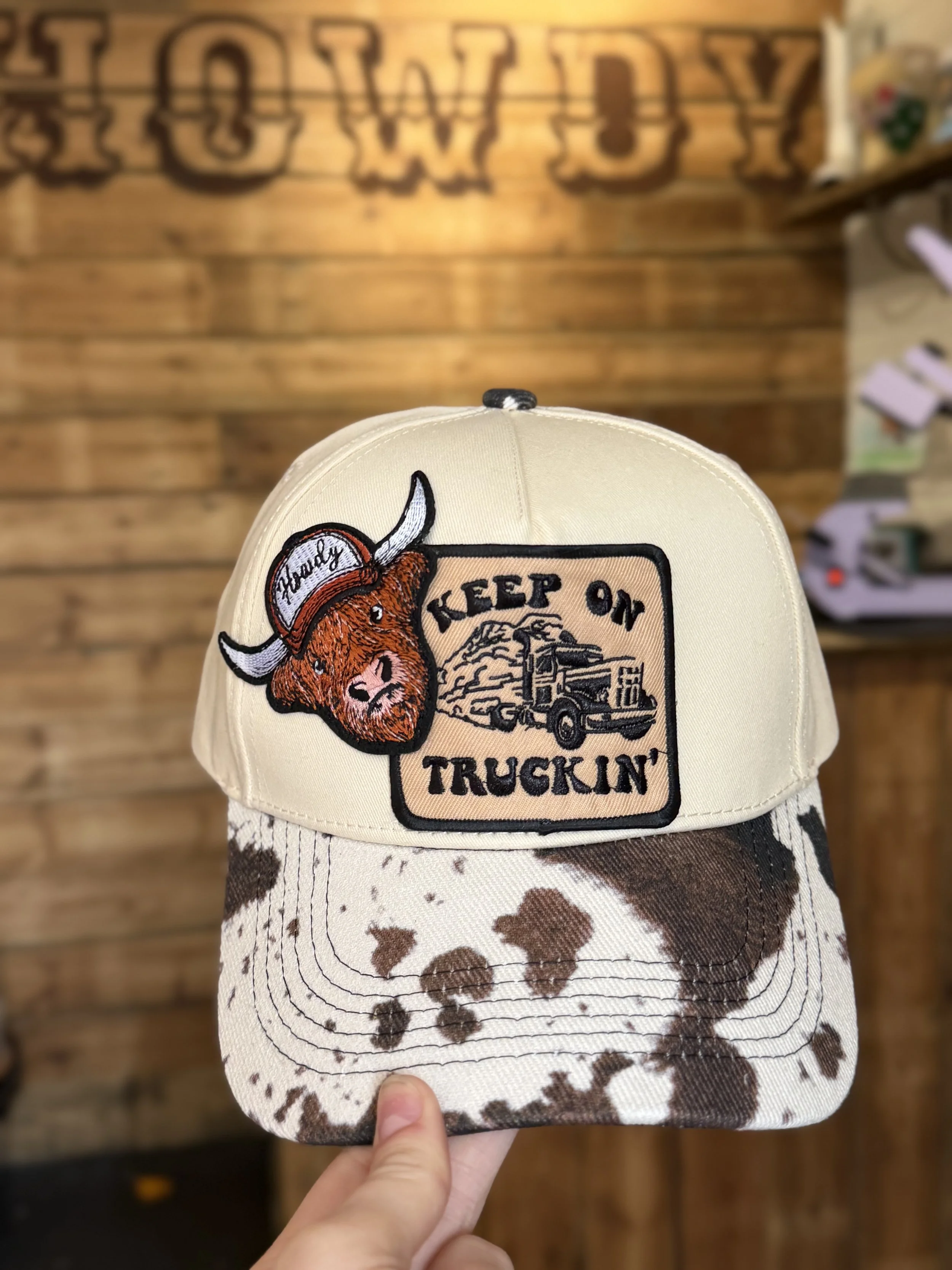 A baseball cap with patches of a bull, a cityscape, and the words 'Keep On Truckin''. The cap has a cow-print brim and is held by a hand against a wooden background.
