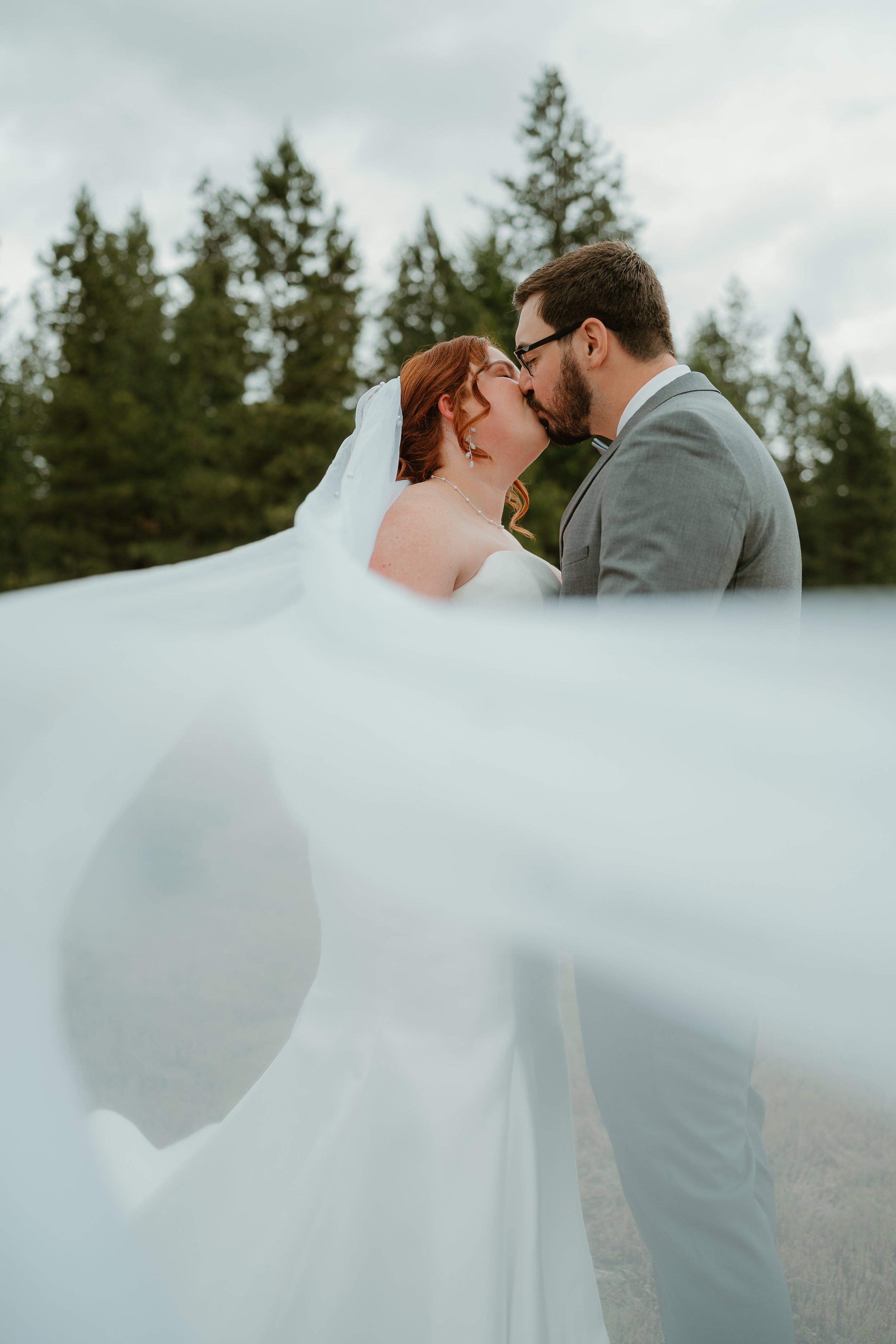 A bride and groom sharing a kiss outdoors, with the bride's veil flowing in the foreground and trees in the background.