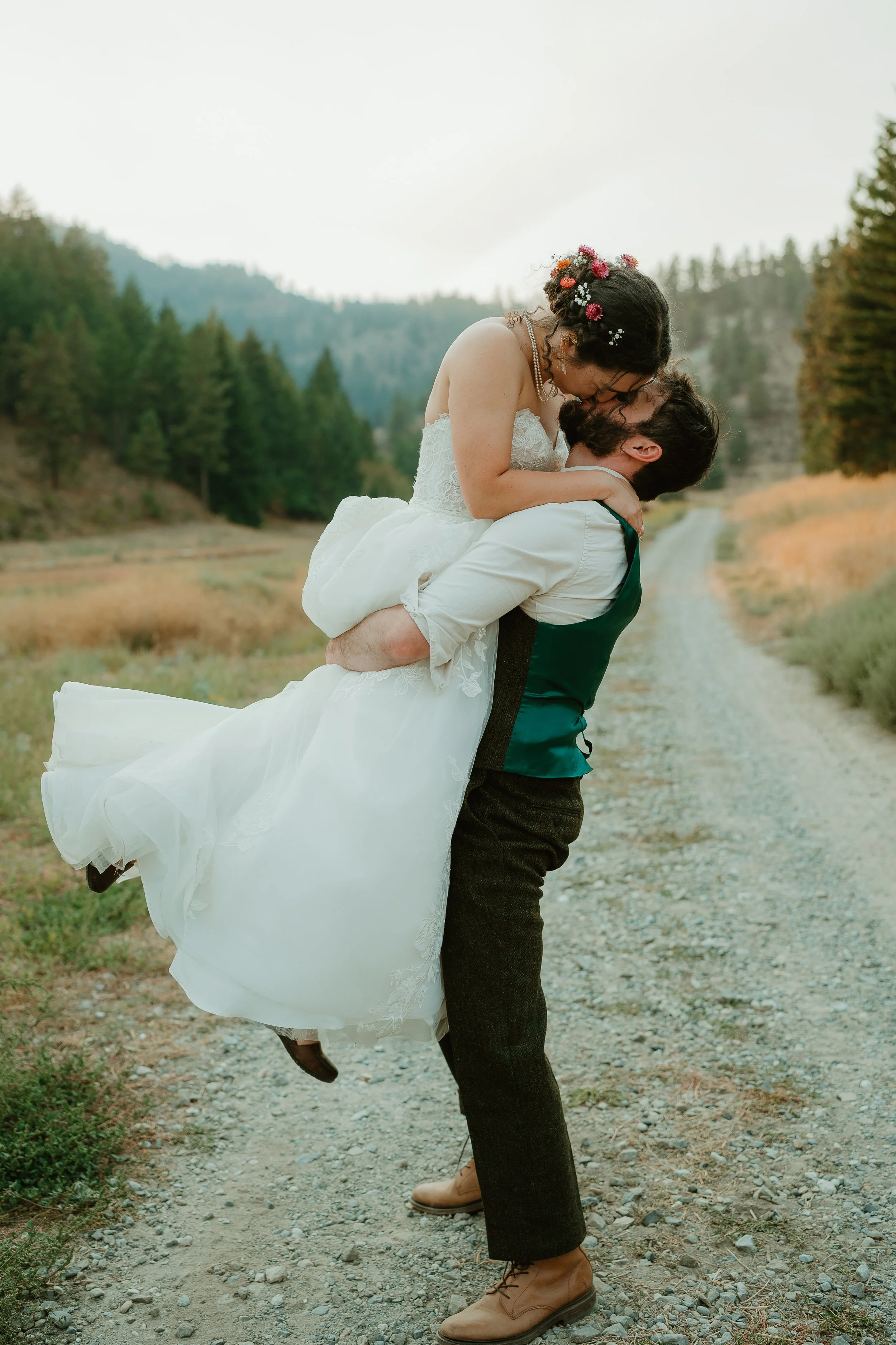A couple in wedding attire sharing a kiss outdoors on a dirt trail surrounded by trees and mountains.