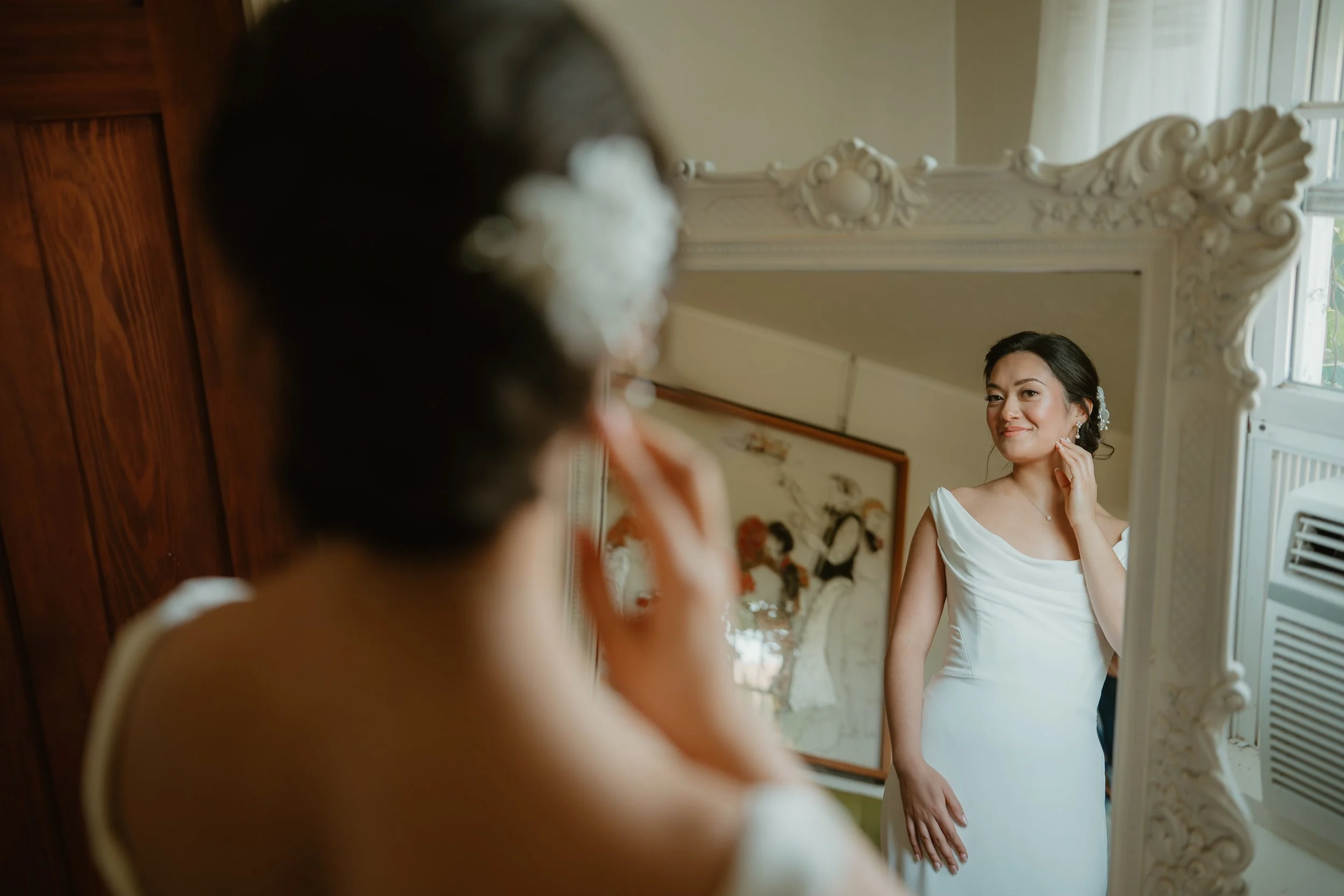 A woman in a white dress looking at herself in an ornate mirror, adjusting her earring.