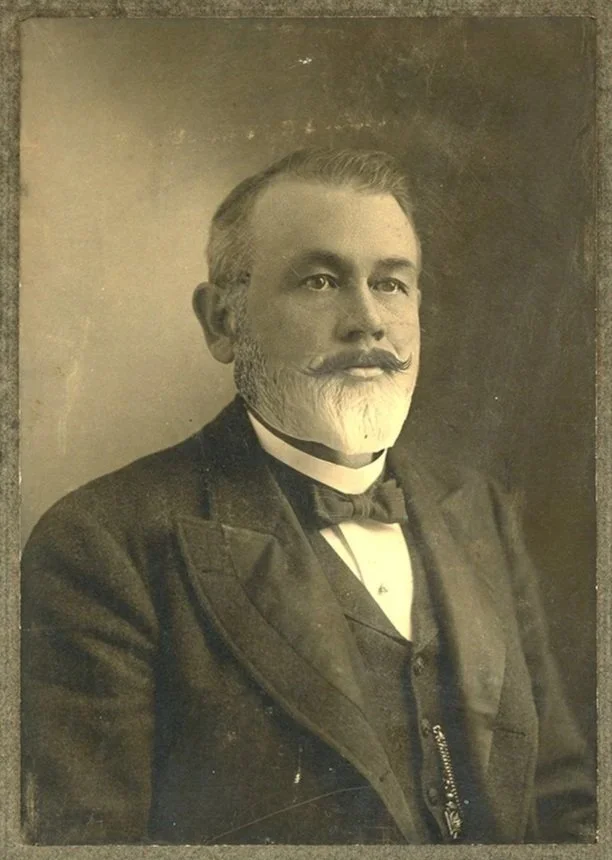 Sepia toned portrait of a man in a dapper suit with short quaffed hair and a grey beard.