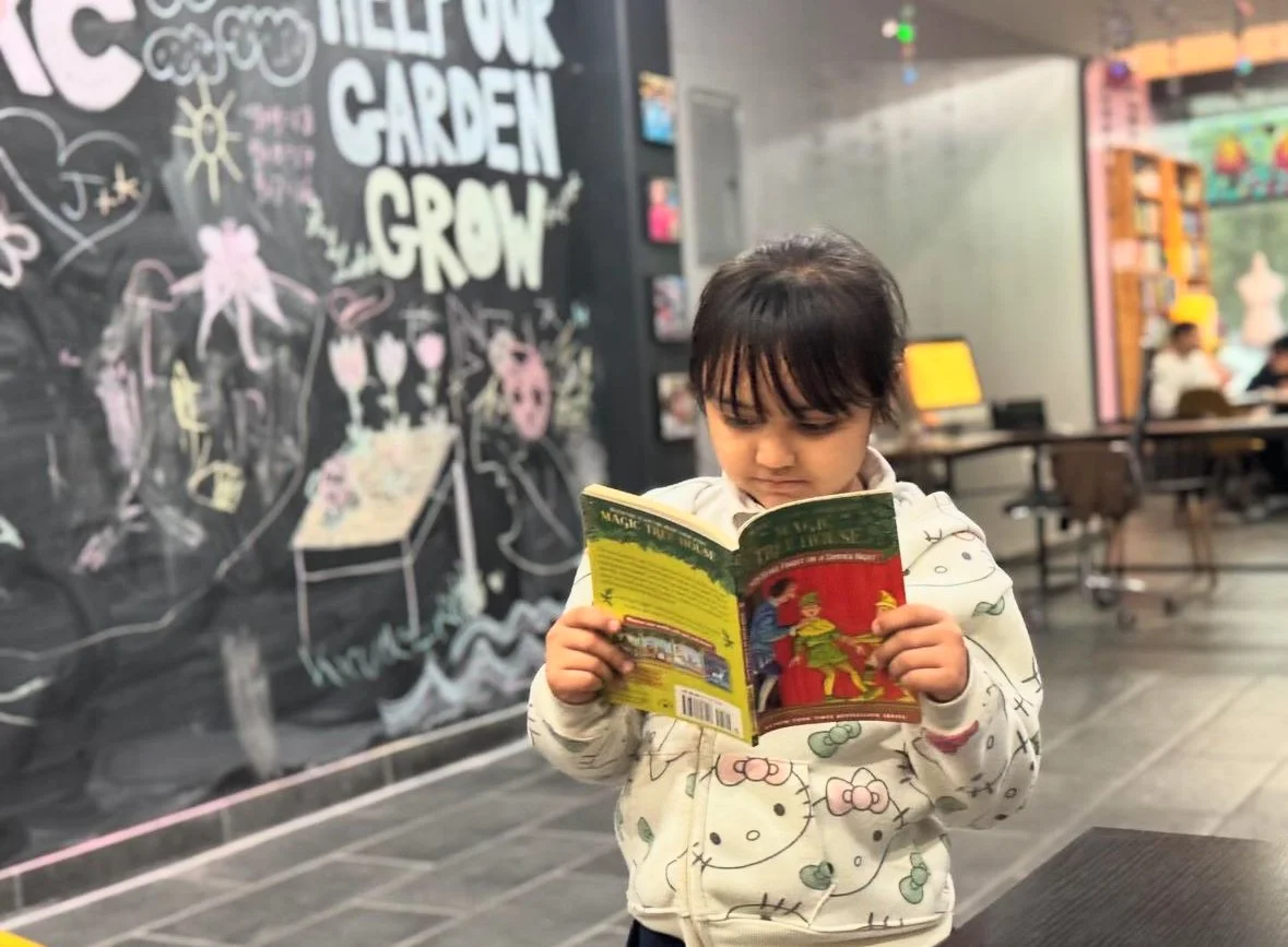 A young girl with dark hair and bangs reading a colorful book indoors, standing in front of a blackboard covered with drawings and writings, with a cafe or library setting visible in the background.