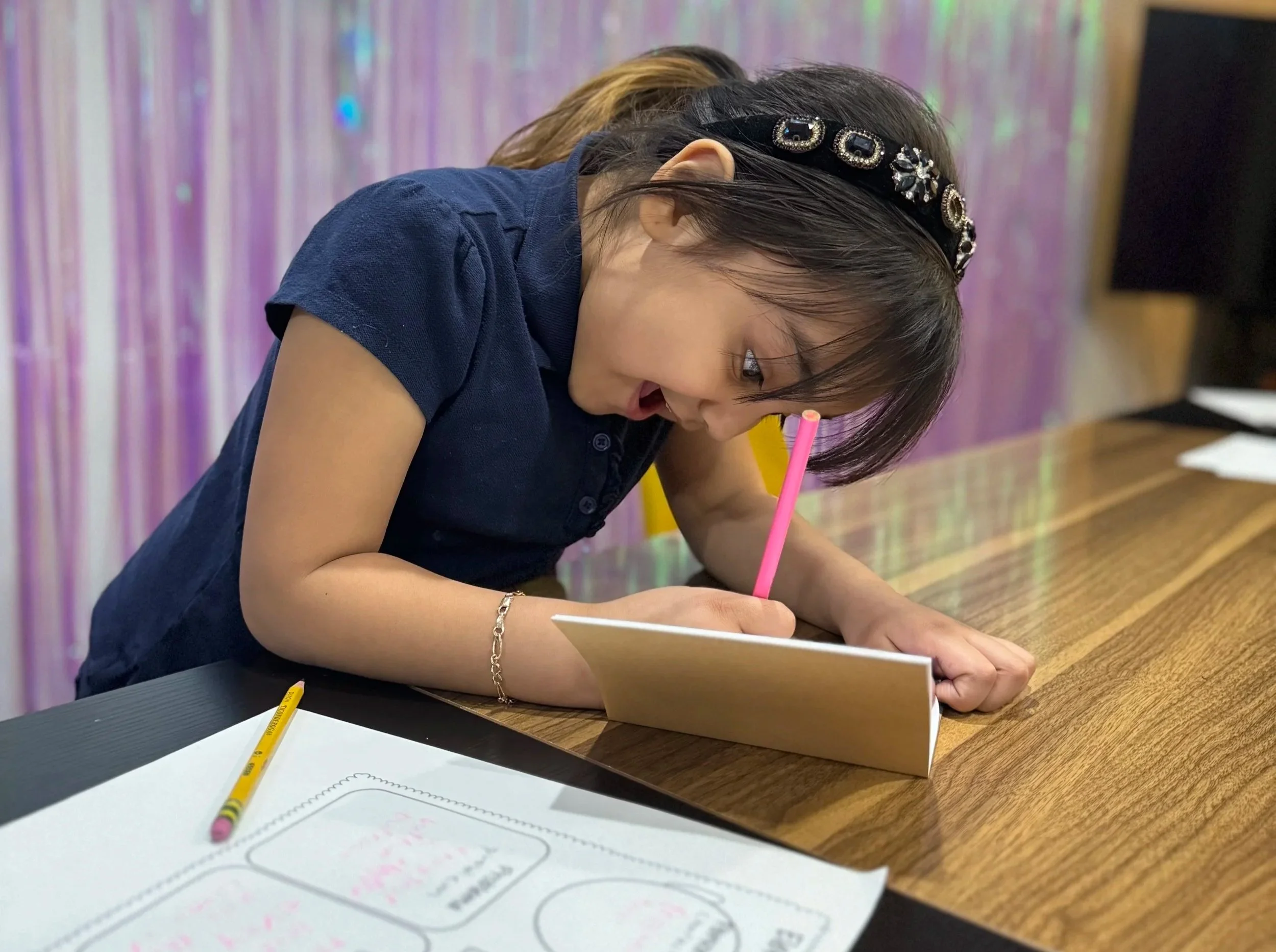 A young girl with dark hair and a black headband is leaning over a wooden table, writing on a notepad with a pink pencil. She is wearing a navy blue shirt, and there are sheets of paper and a yellow pencil on the table.
