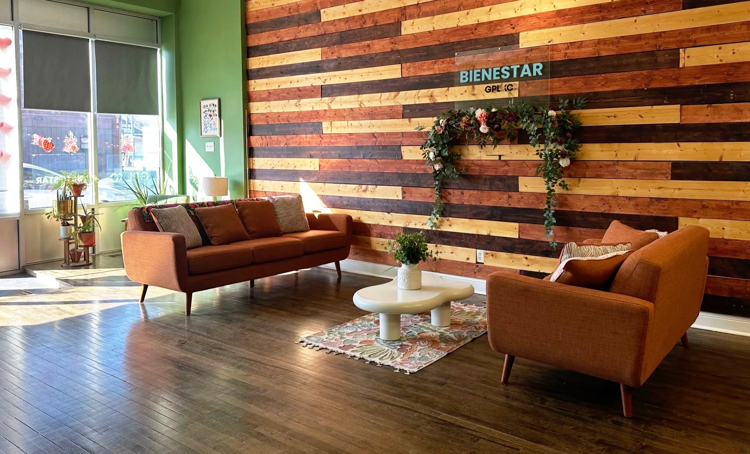 A cozy waiting area with two mid-century modern sofas in warm earth tones, a white coffee table with a potted plant, a colorful rug, and a wooden accent wall with the word 'BIENESTAR' and decorated with a floral wreath. Natural light streams through large windows.