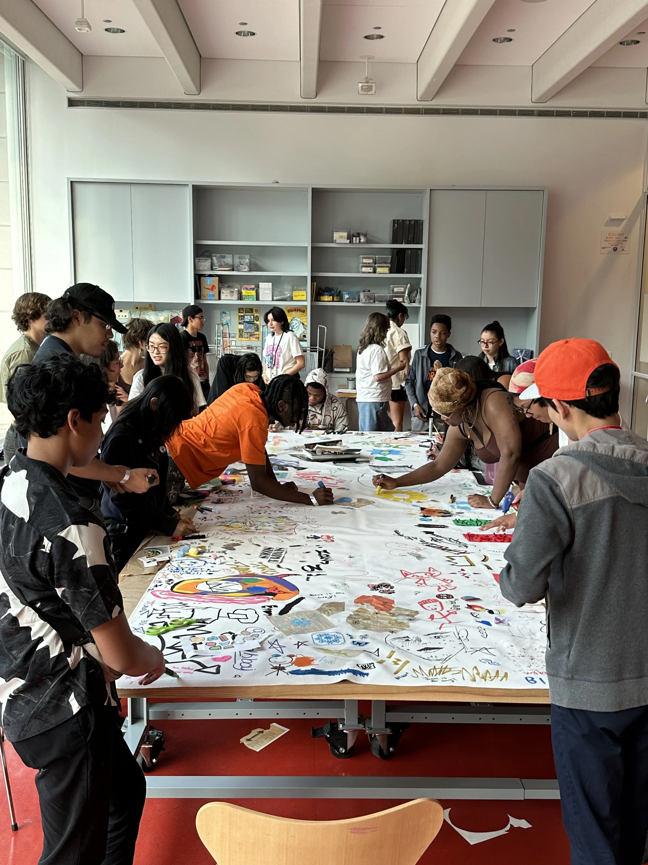 People gathered around a large table, creating and drawing on a large sheet of paper with various colorful designs and messages, in a room with shelves and windows.