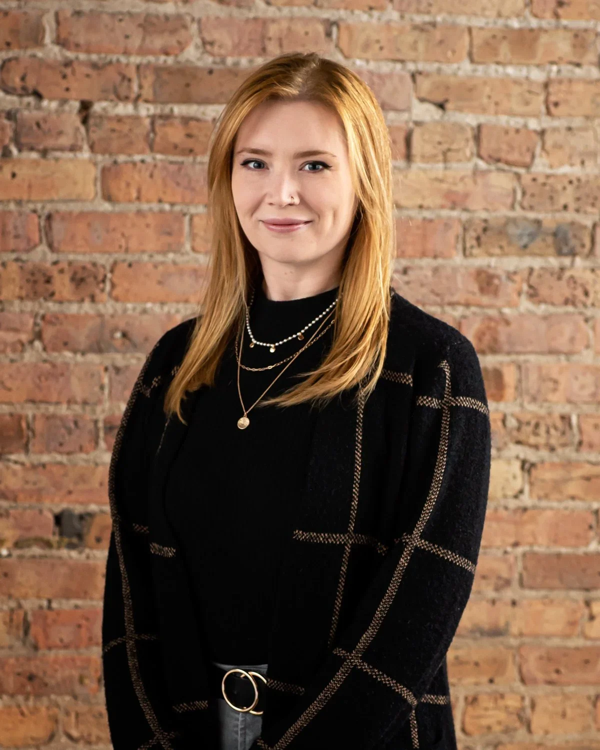 A woman with red hair, wearing a black top, layered necklaces, and a black blazer with a checkered pattern, standing against an exposed brick wall.