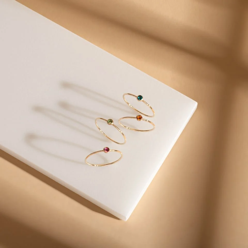 Four gold rings with small colored gemstones on a white surface with dramatic lighting and long shadows.