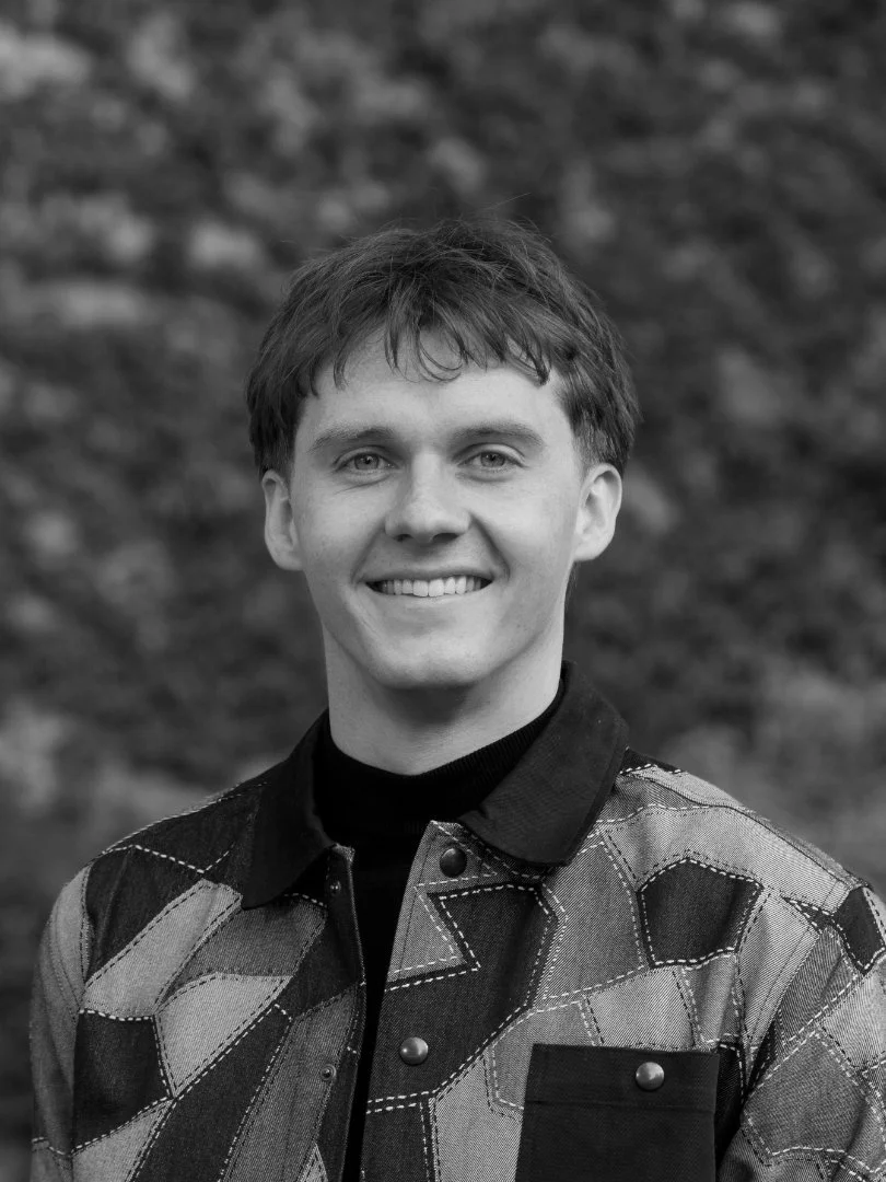 Black and white portrait of a young man smiling outdoors, wearing a patterned jacket over a dark shirt, with a blurred natural background.