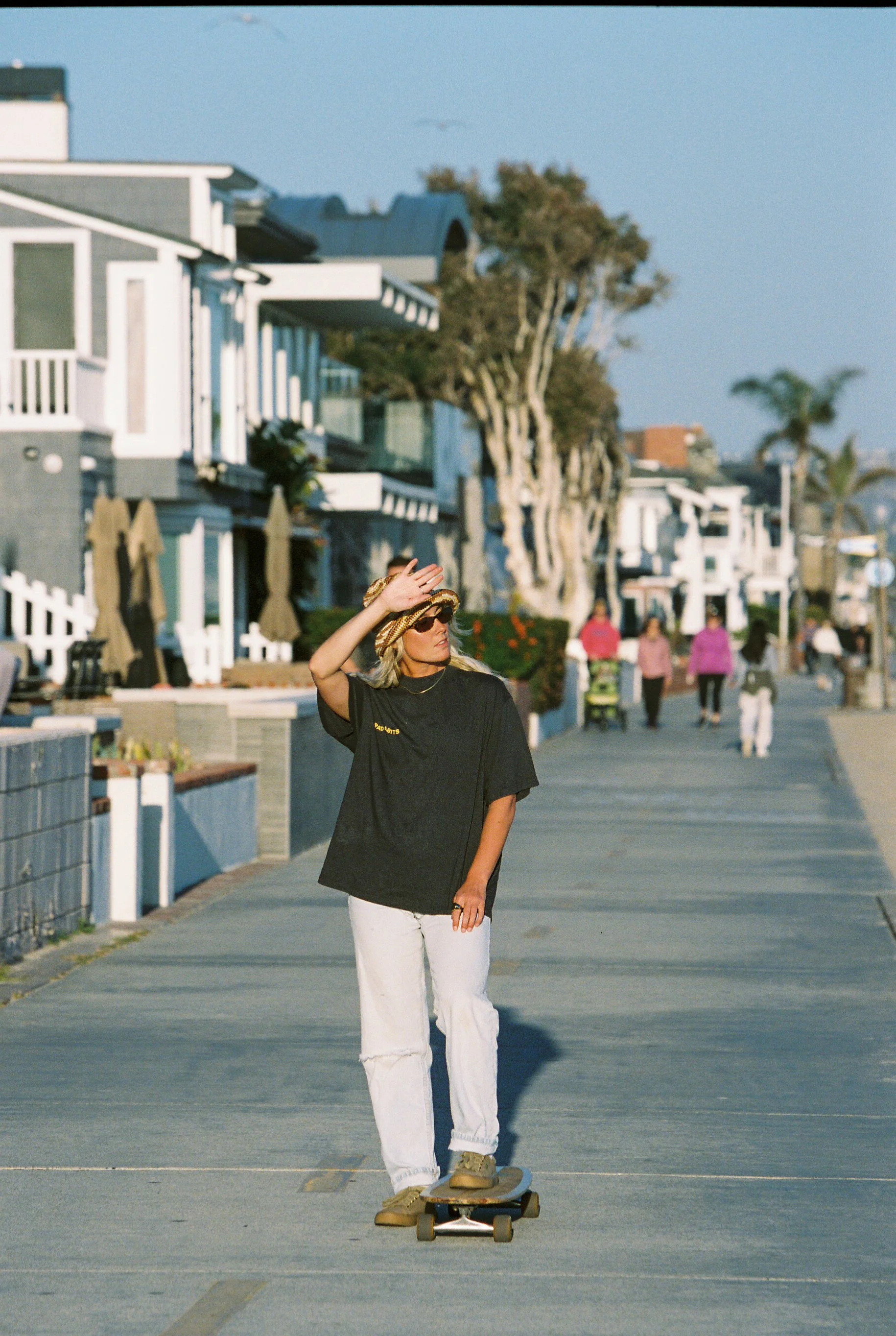 Rad Habits Charcoal Tee worn by a woman skateboarding on a sidewalk in Newport Beach with beach houses and palm trees in the background.