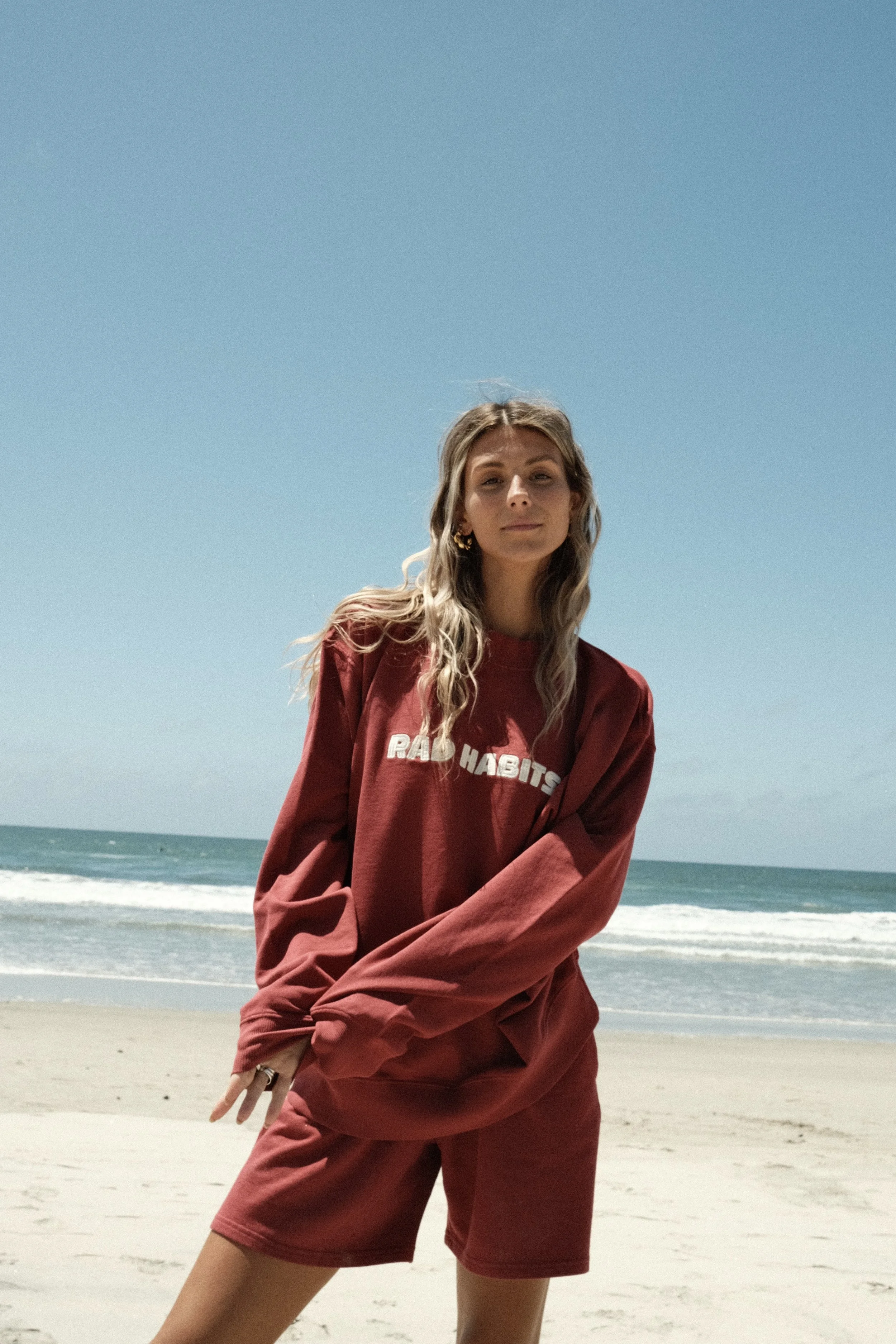Red Rad Habits 100% organic crewneck and 100% organic sweatshorts. Image of a young woman in a red crewneck and shorts standing on a sandy beach with the ocean and blue sky in the background.