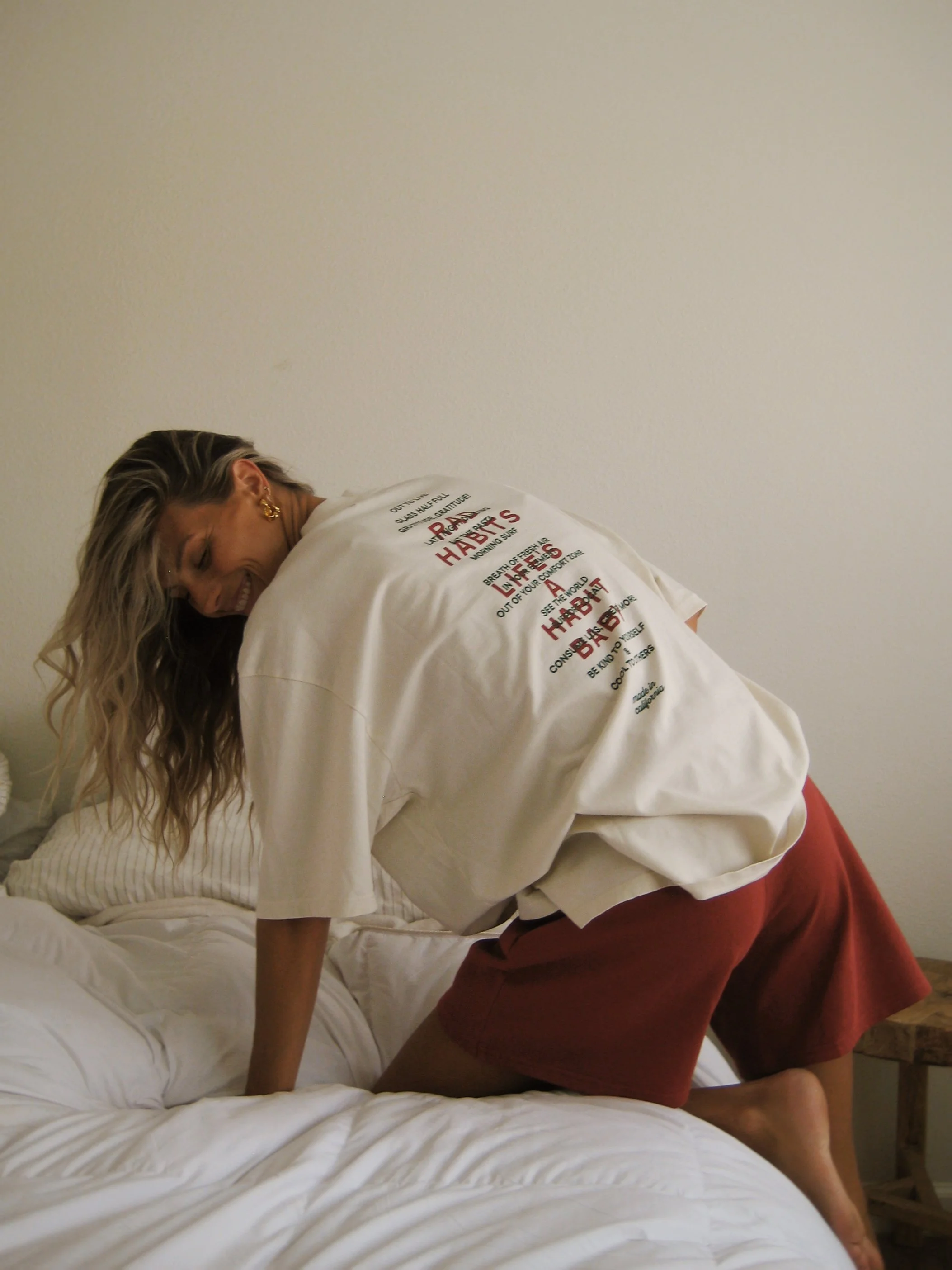 A woman with long wavy hair, wearing a white T-shirt and red shorts, smiling as she leans over a bed. Wearing Rad Habits Tee and red 100% organic cotton sweat shorts.