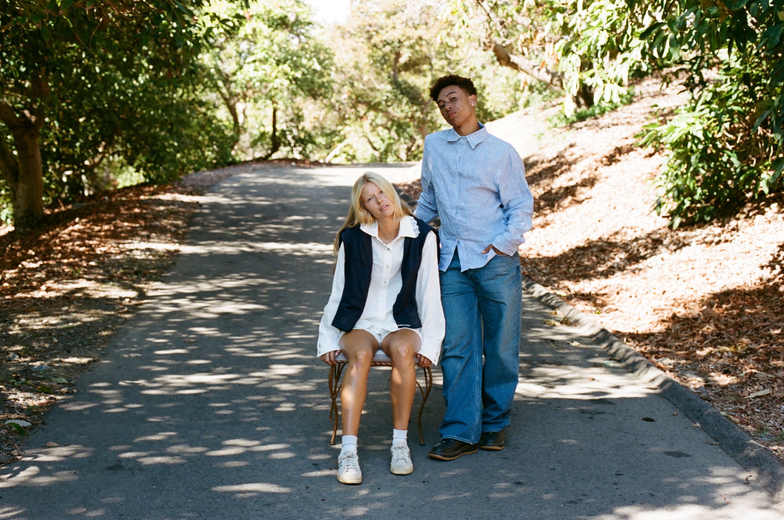 A young woman with blonde hair seated on a wooden chair on a shaded outdoor path, with a young man standing beside her, both dressed casually in light-colored clothing, surrounded by greenery.