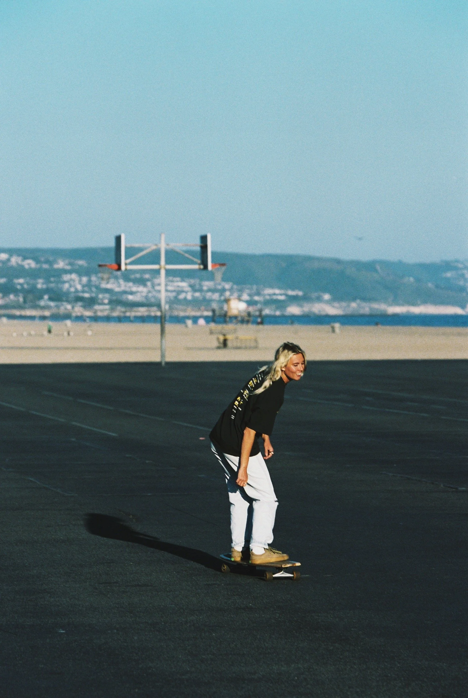 A young woman skateboarding on a black asphalt surface near a beach with mountains in the background, under a clear blue sky.