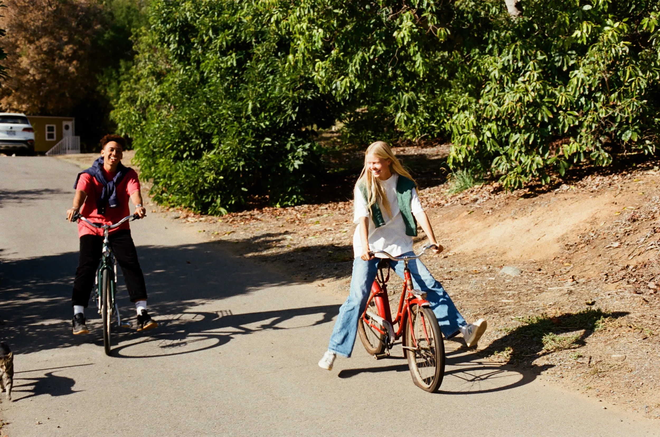 Two children riding bikes on a paved road surrounded by greenery and trees during daytime, smiling and enjoying the outdoor activity.