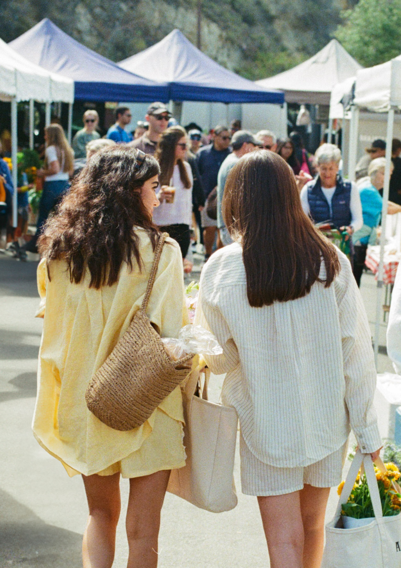 Two women shopping at an outdoor market, viewed from behind. One woman has dark, curly hair and a large woven bag, while the other has straight, brown hair and a white tote. Tents and other shoppers are in the background.