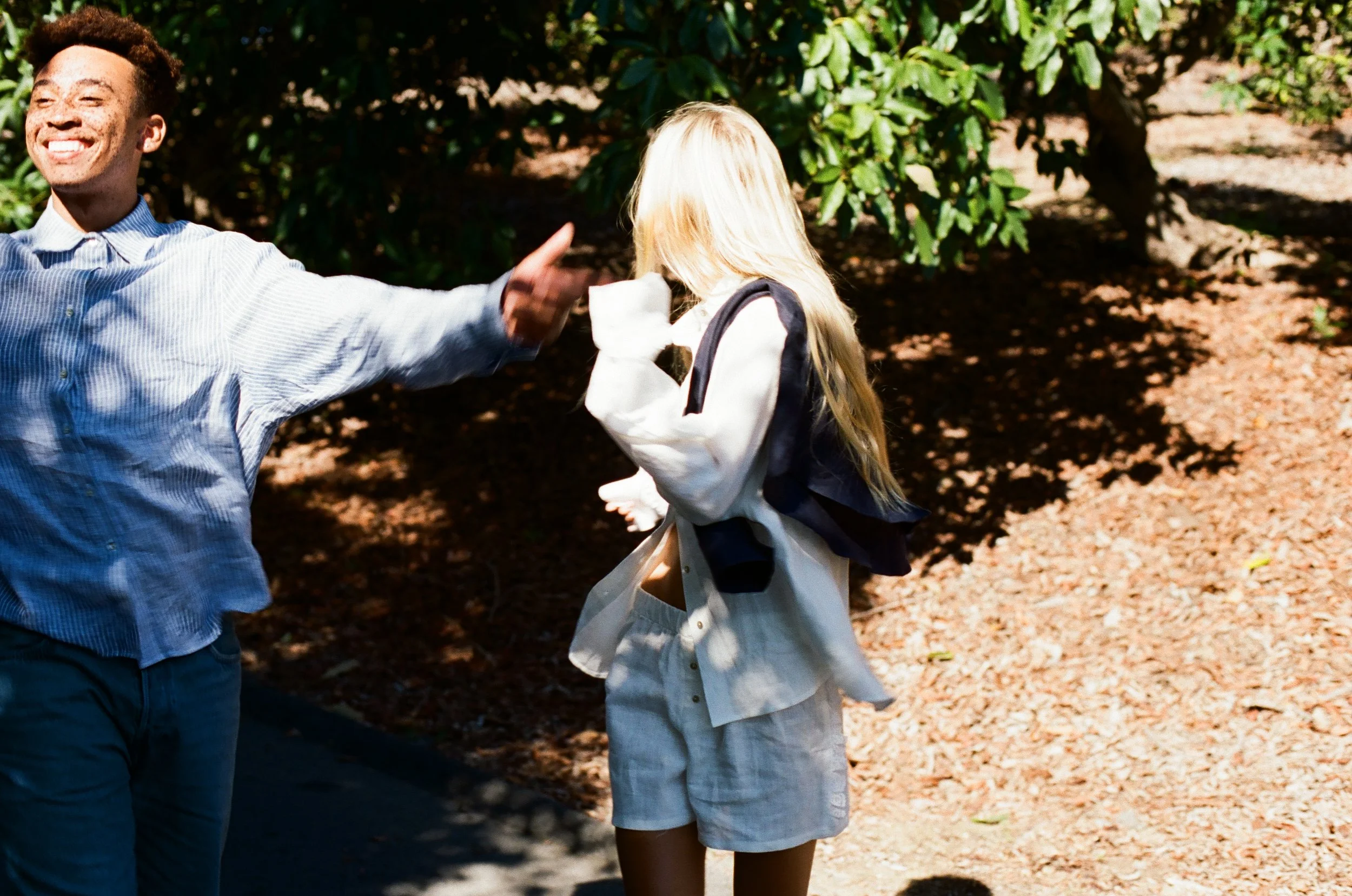 A man is smiling and performing a high five with a blonde girl, who is turned away, on a sunny outdoor pathway surrounded by greenery.