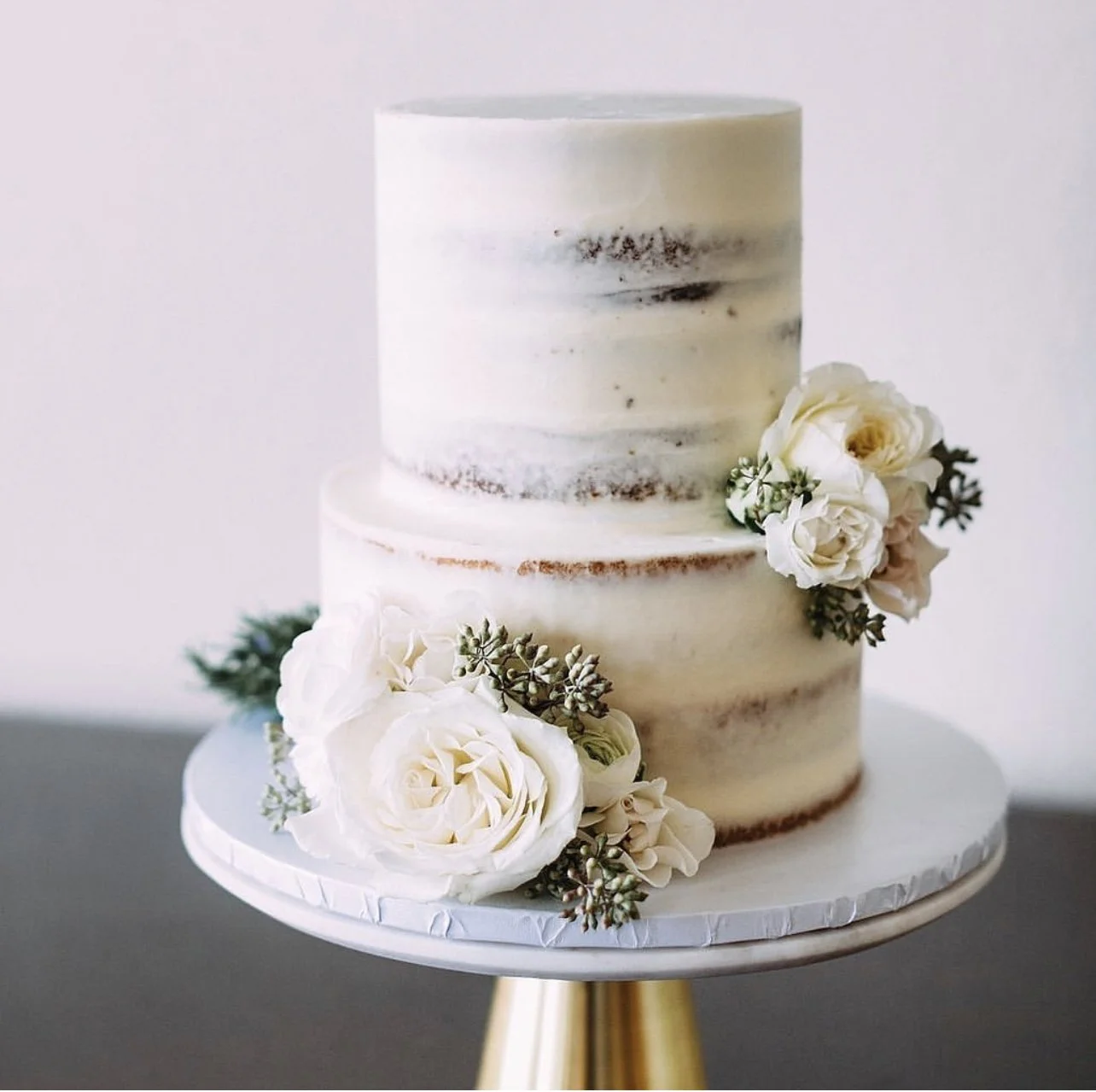 Two-tiered naked wedding cake with white and cream frosting, decorated with white roses and greenery, on a gold stand.