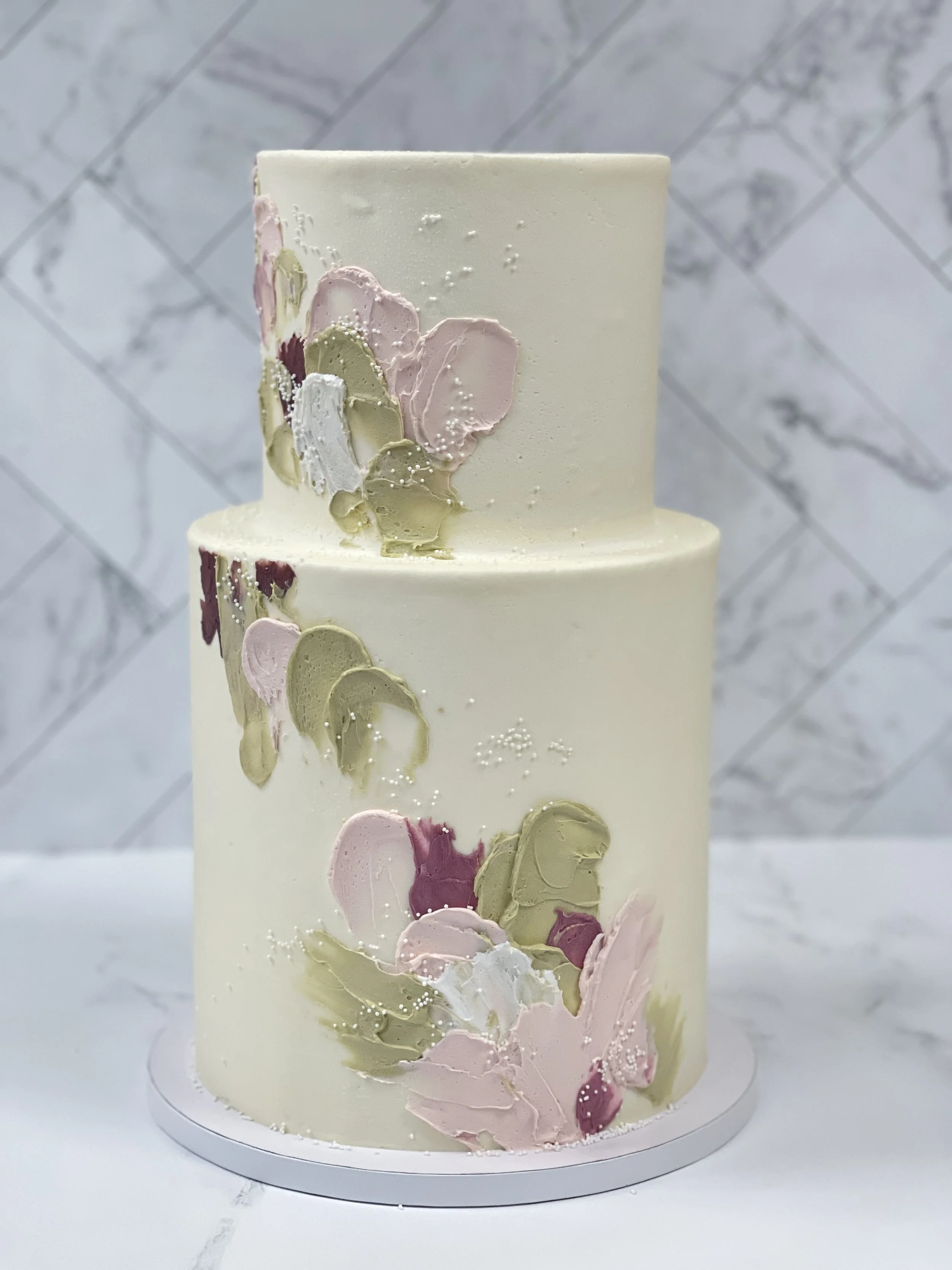 Two-tiered white cake decorated with pastel-colored abstract floral icing in pink, green, white, and burgundy, placed on a white marble surface with a marble wall background.