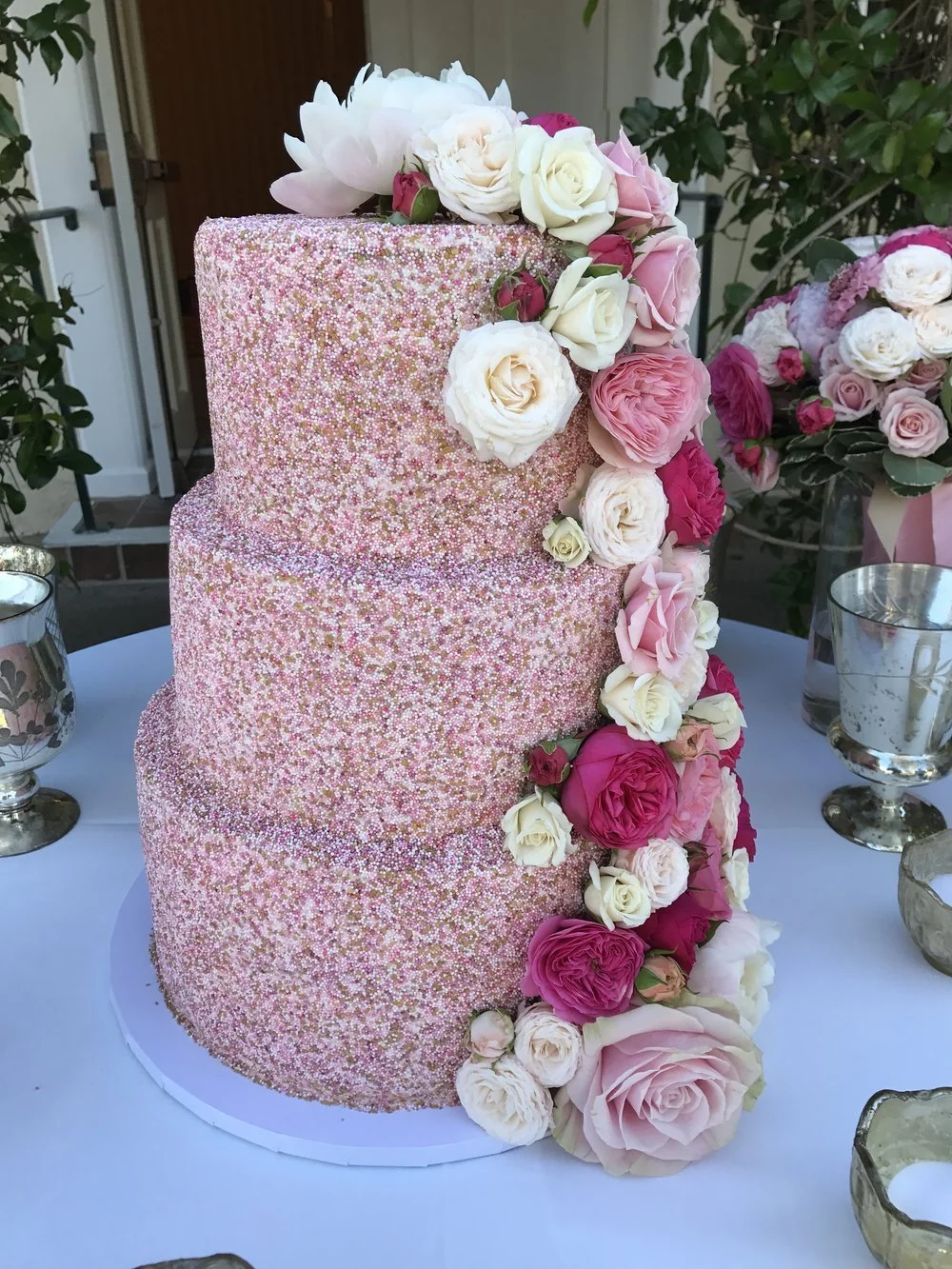 Three-tier pink wedding cake decorated with small multicolored sprinkles and cascading pink, white, and cream roses on a white cake board, with silver candle holders and pink floral arrangements in the background.