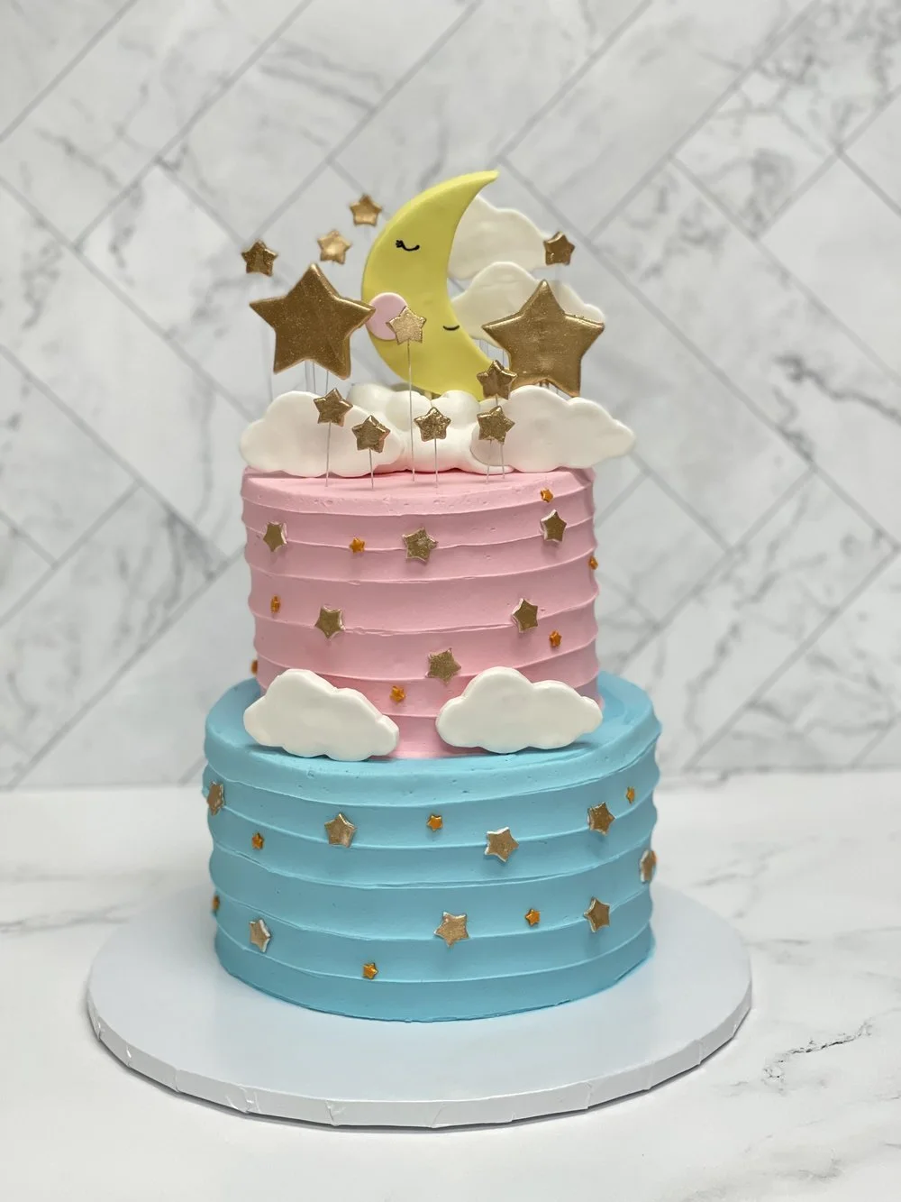 Two-tiered birthday cake with pastel pink and blue frosting, decorated with white clouds and gold star sprinkles. The cake top features a crescent moon with closed eyes, small clouds, and gold star decorations, all set against a marble tile background.