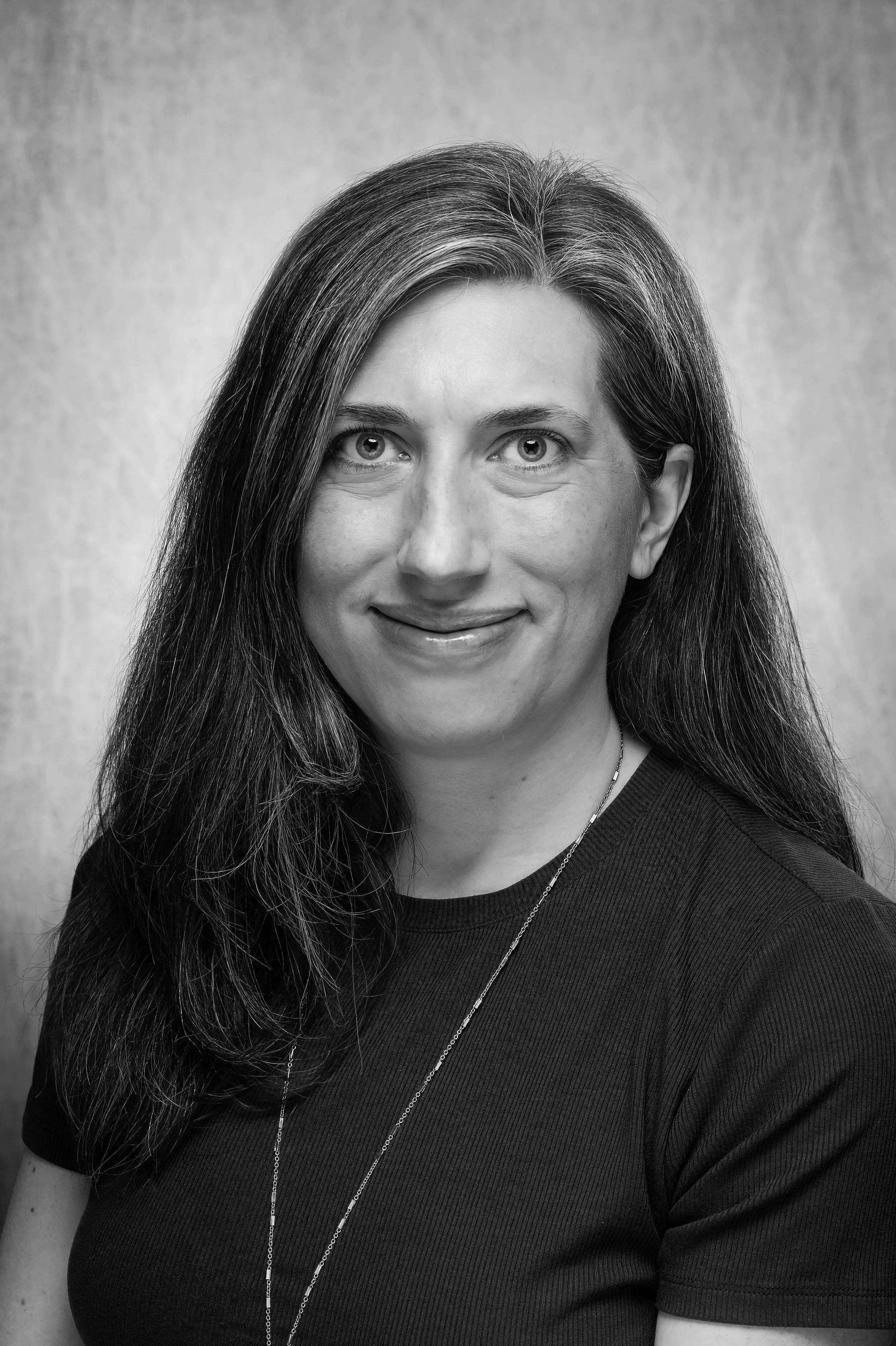 Black and white portrait of a woman with long hair, wearing a dark top, necklace, and smiling slightly, against a plain background.