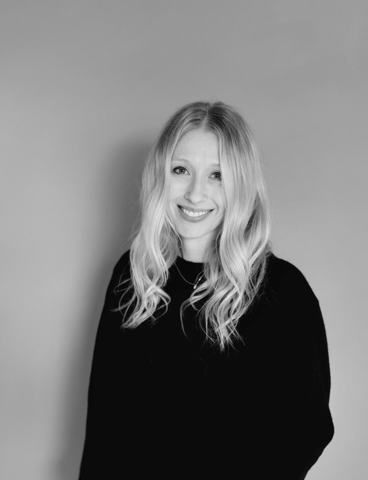 A smiling woman with long, wavy blonde hair, wearing a black top, against a plain background.