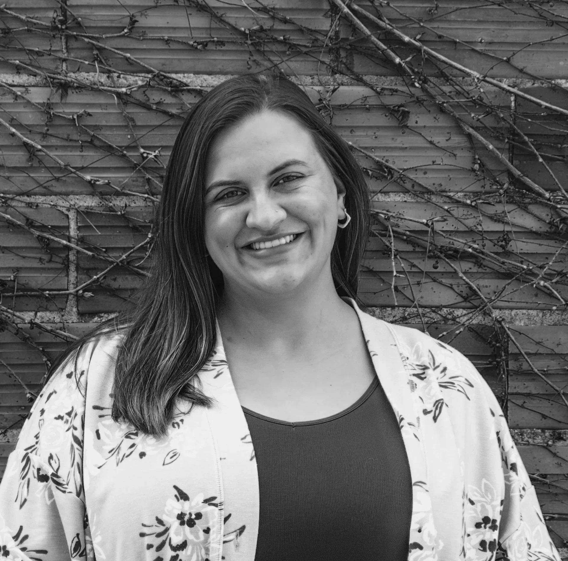 Black and white photo of a smiling woman with long hair, wearing a patterned jacket and a dark top, standing in front of a wooden wall with vines.