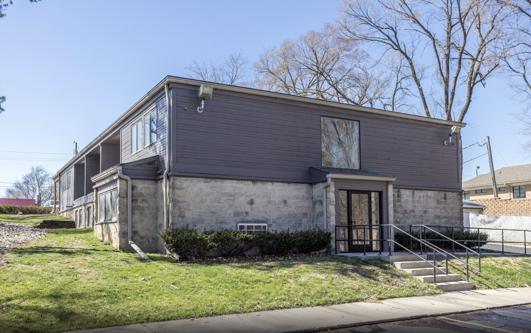 Two-story building with a stone foundation and dark siding, with stairs and a ramp leading to the entrance, and trees with leafless branches in the background.